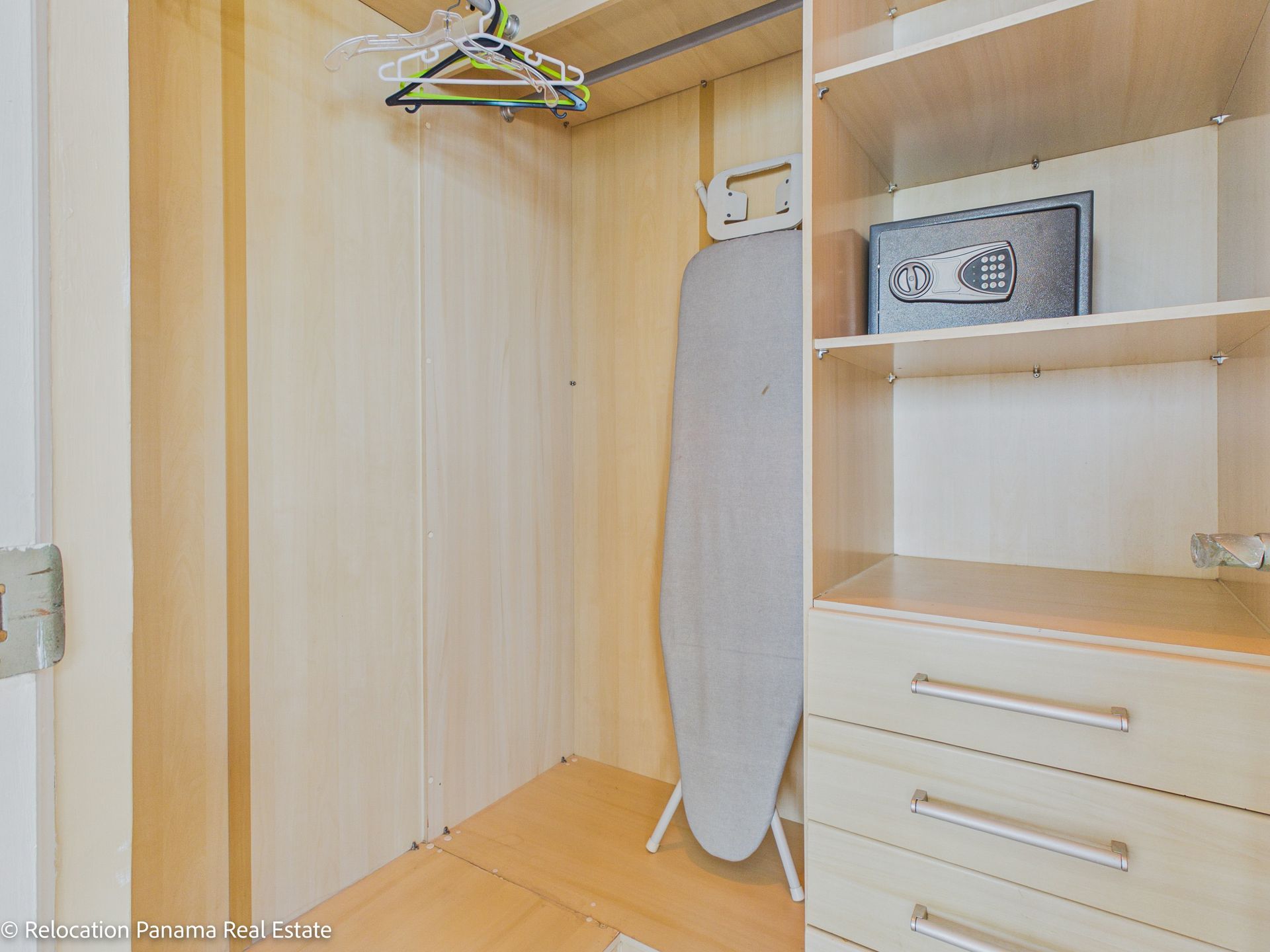 Un armario de madera con perchero, tabla de planchar de pie, estantes abiertos y una caja fuerte electrónica gris empotrada en la pared.
