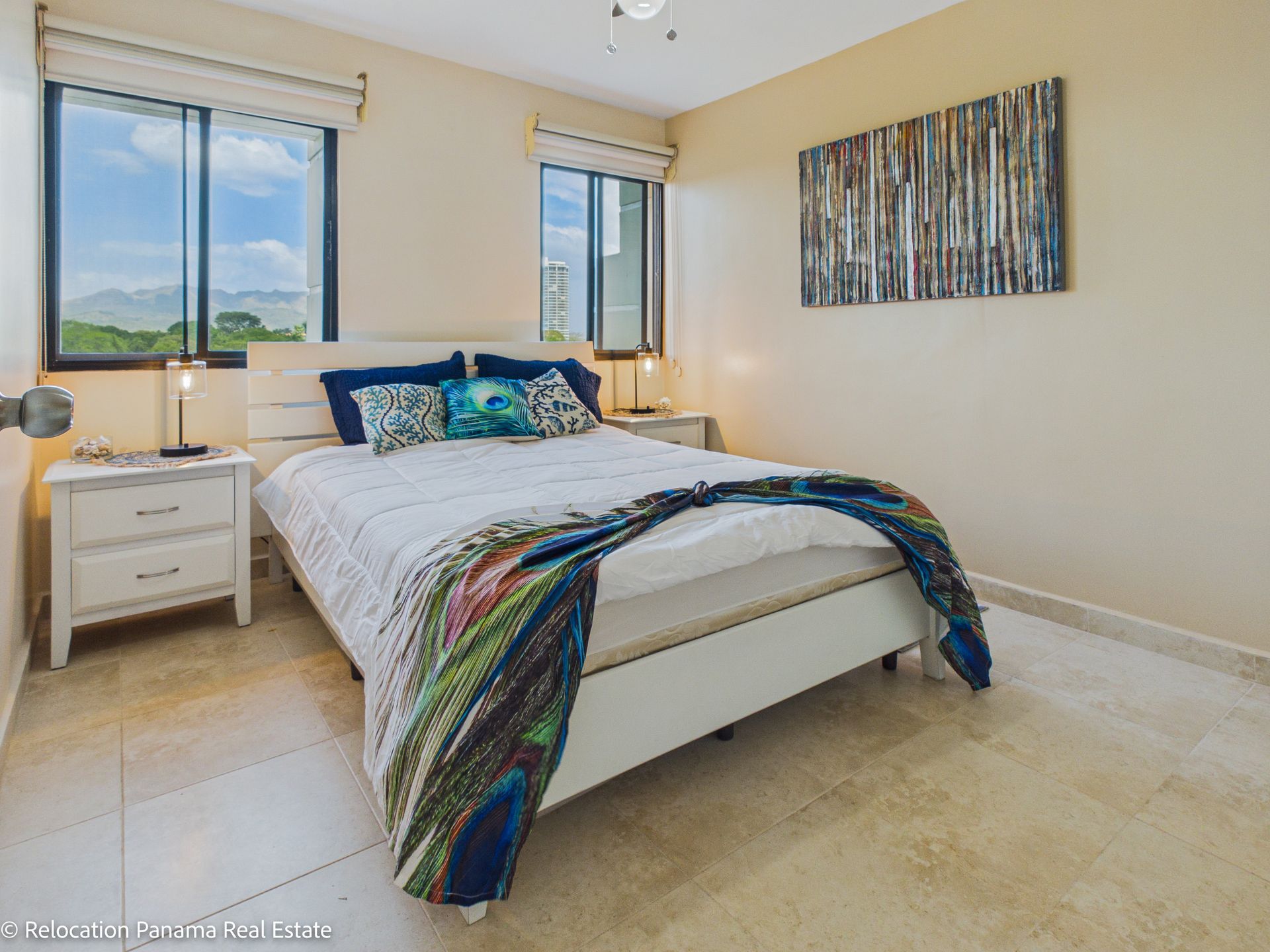 Un dormitorio luminoso con una cama, una mesita de noche blanca, dos ventanas con vistas a las montañas y una obra de arte abstracto en una pared de color beige.