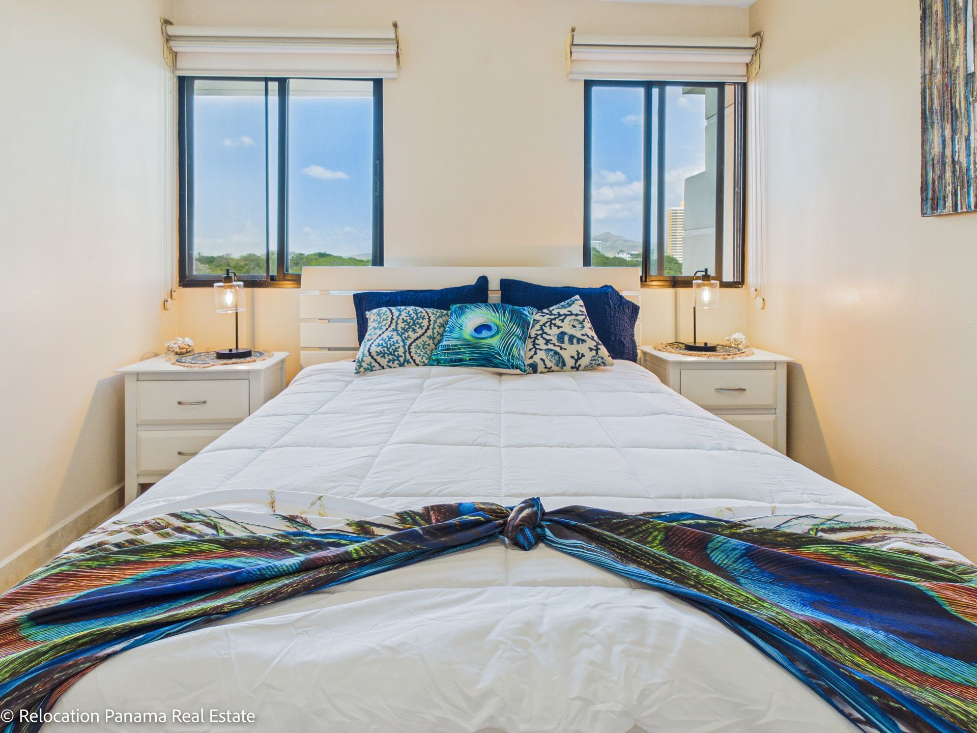 Un dormitorio luminoso con una cama de matrimonio situada entre dos ventanas y mesitas de noche blancas, coronadas con cojines estampados.