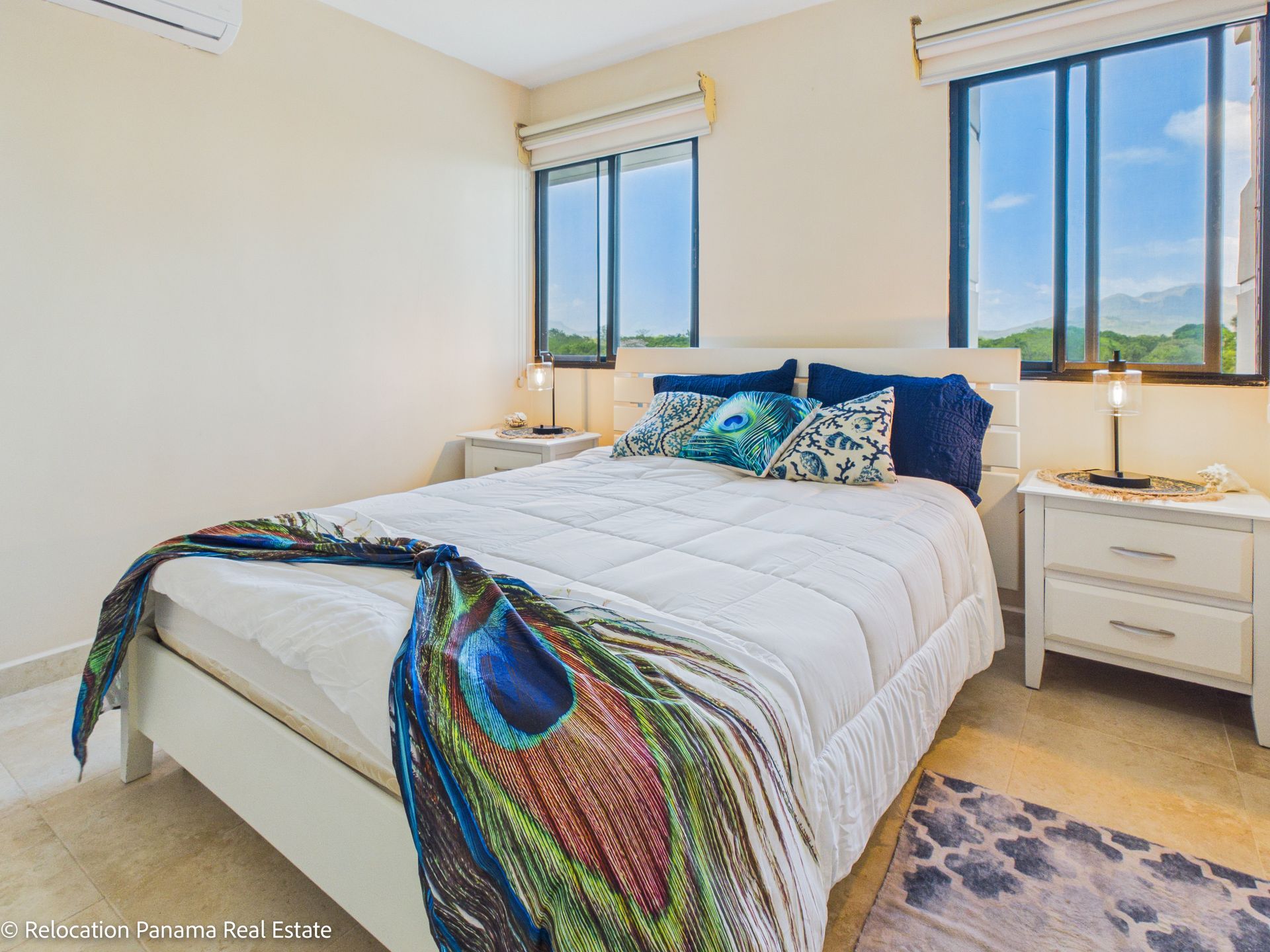 Un dormitorio blanco con una cama con una manta estampada con plumas de pavo real, flanqueado por dos mesitas de noche y dos ventanas.