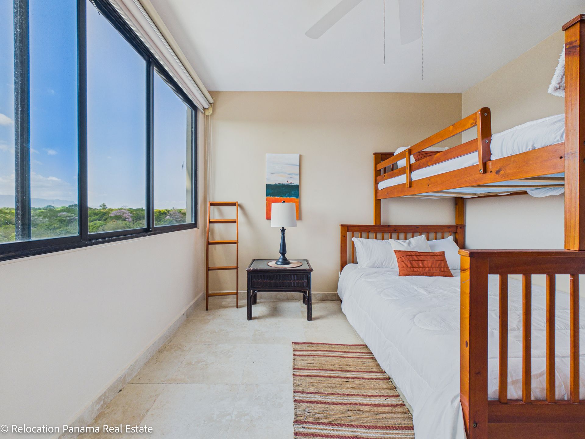 Un dormitorio con una litera de madera, una pequeña mesita de noche negra con una lámpara y una gran ventana con vistas a la vegetación.