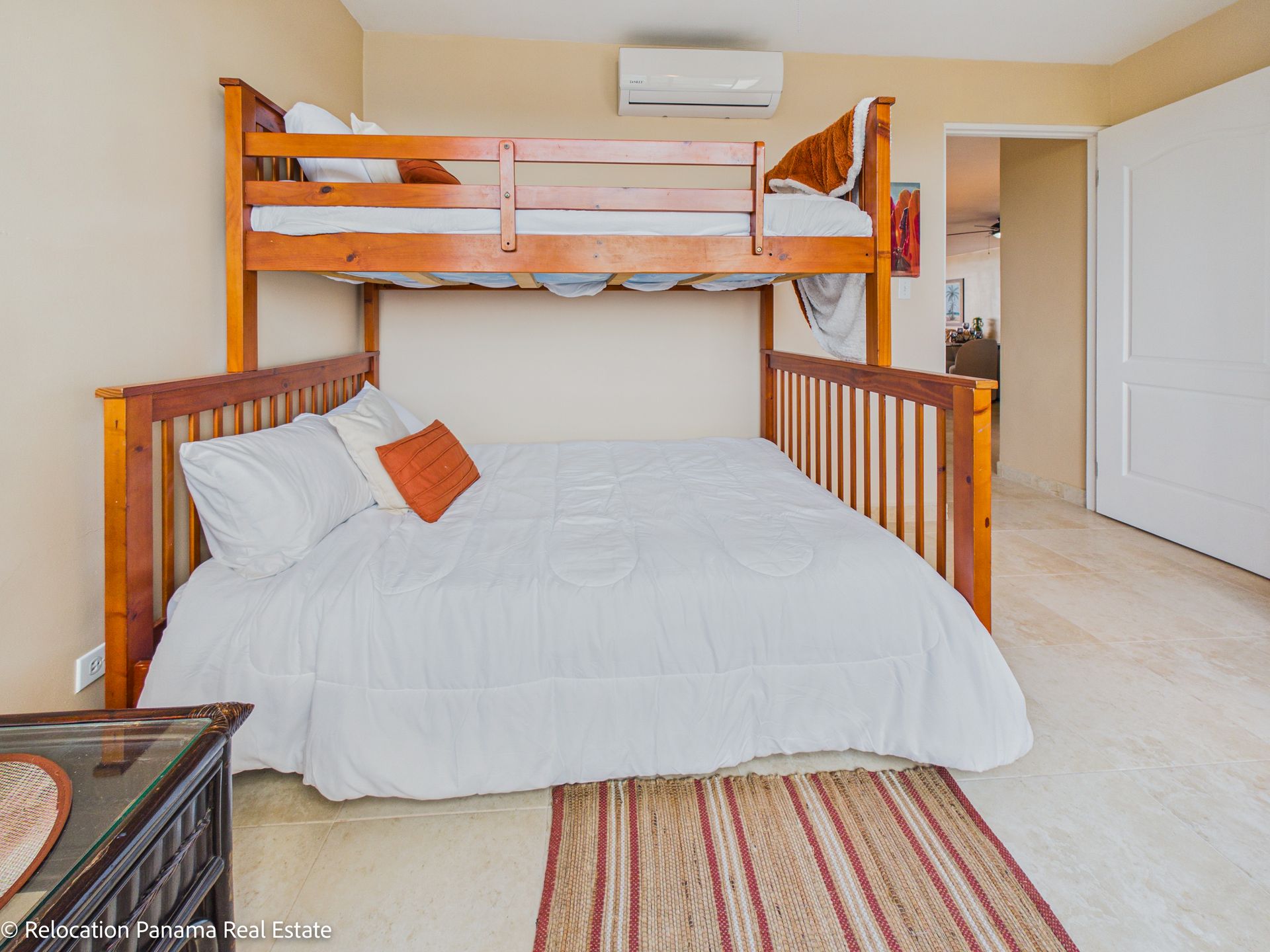 Una litera de madera con una cama inferior grande, cubierta con un edredón blanco y un cojín decorativo naranja, en un dormitorio sencillo.