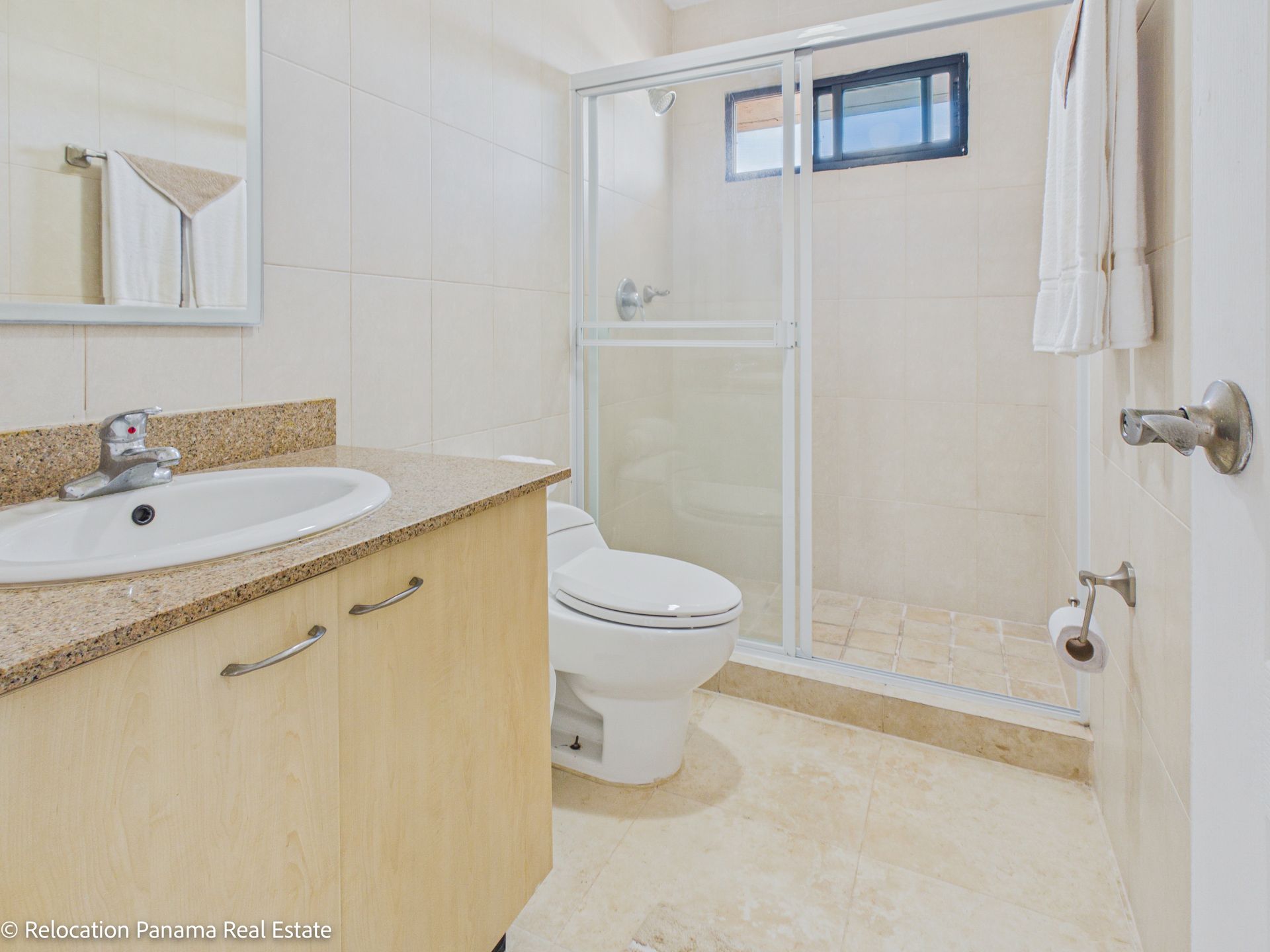Un baño en tonos neutros que cuenta con un lavabo con tocador, un inodoro y una cabina de ducha con mampara de cristal y una pequeña ventana.