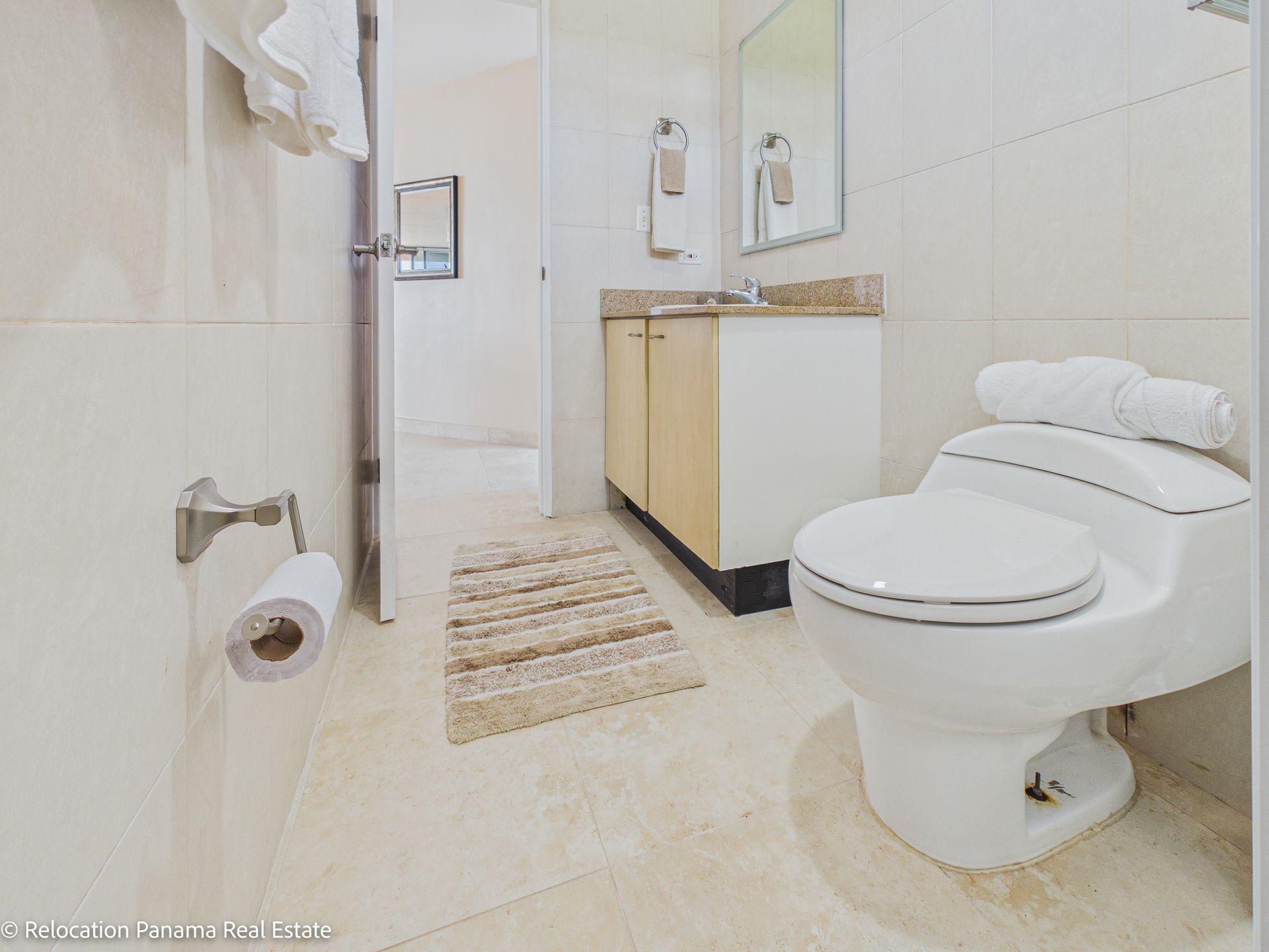 Un baño en tonos neutros con inodoro blanco, tocador de madera clara con lavabo, alfombra beige y portarrollos de papel higiénico.