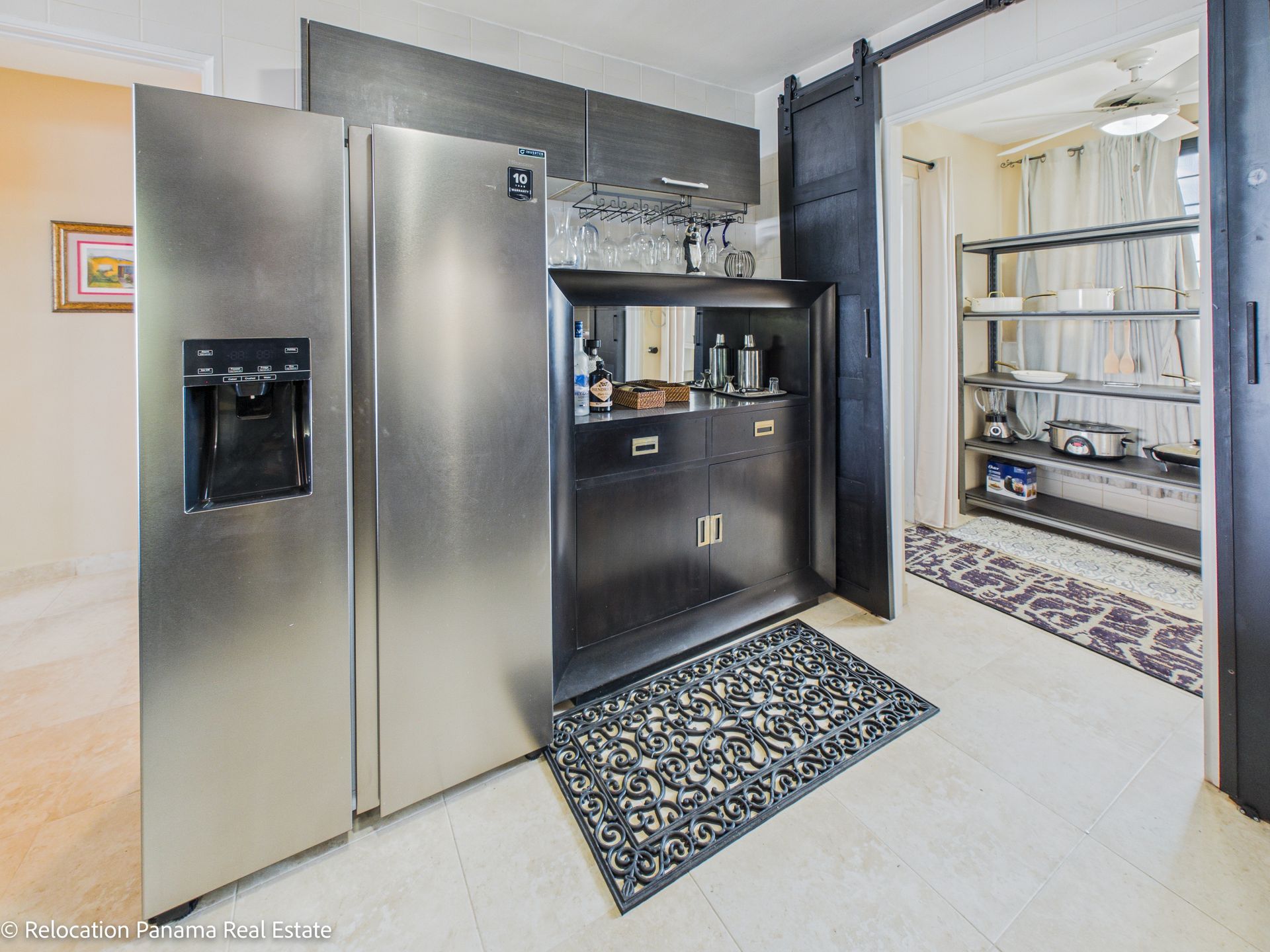 Un refrigerador de acero inoxidable junto a un mueble bar negro con espejo y estantes para vasos, y una despensa en la parte trasera.