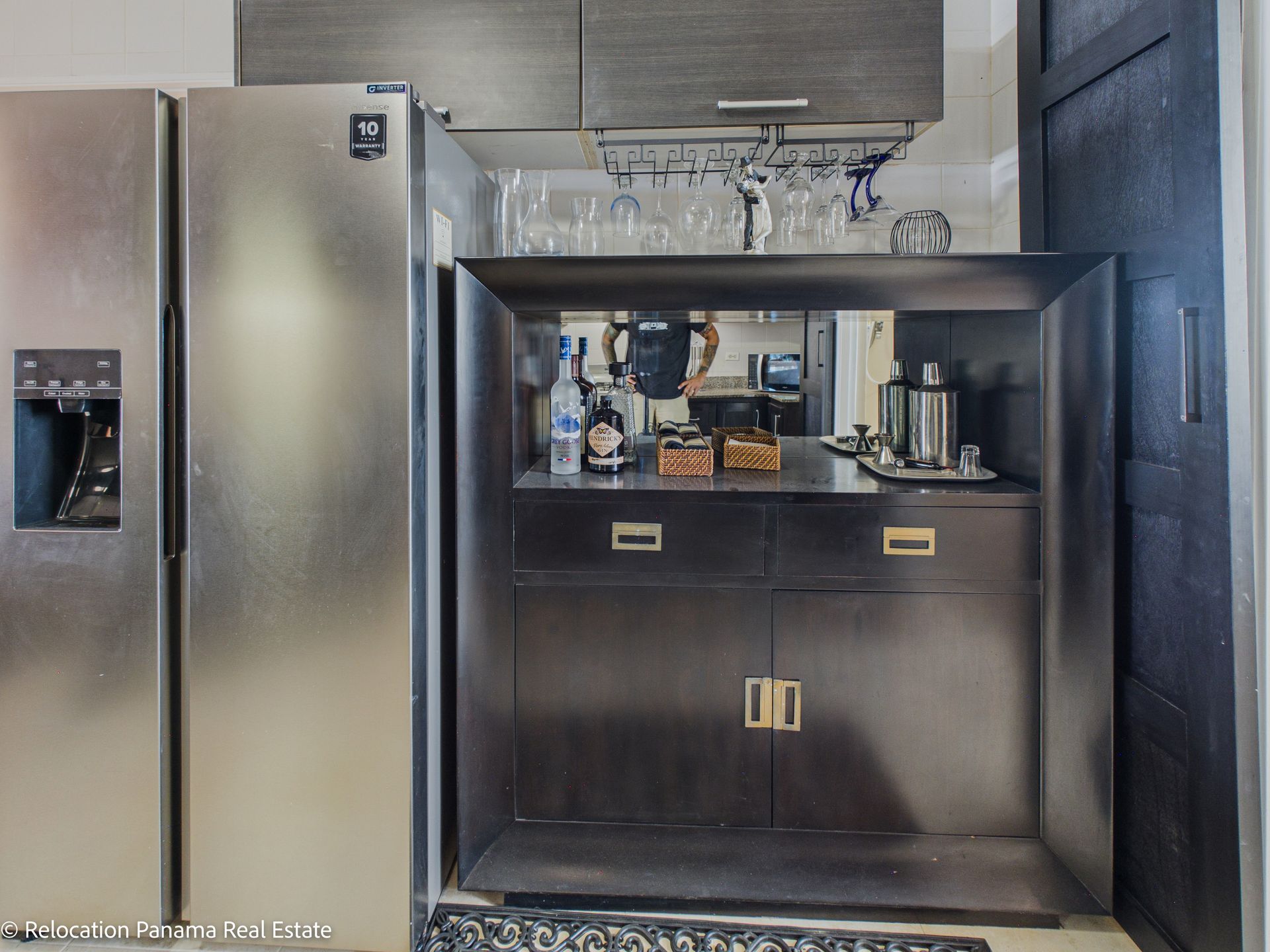 Un frigorífico de acero inoxidable se encuentra junto a una barra de bar de madera oscura con cristalería, botellas y una pared trasera con espejo.