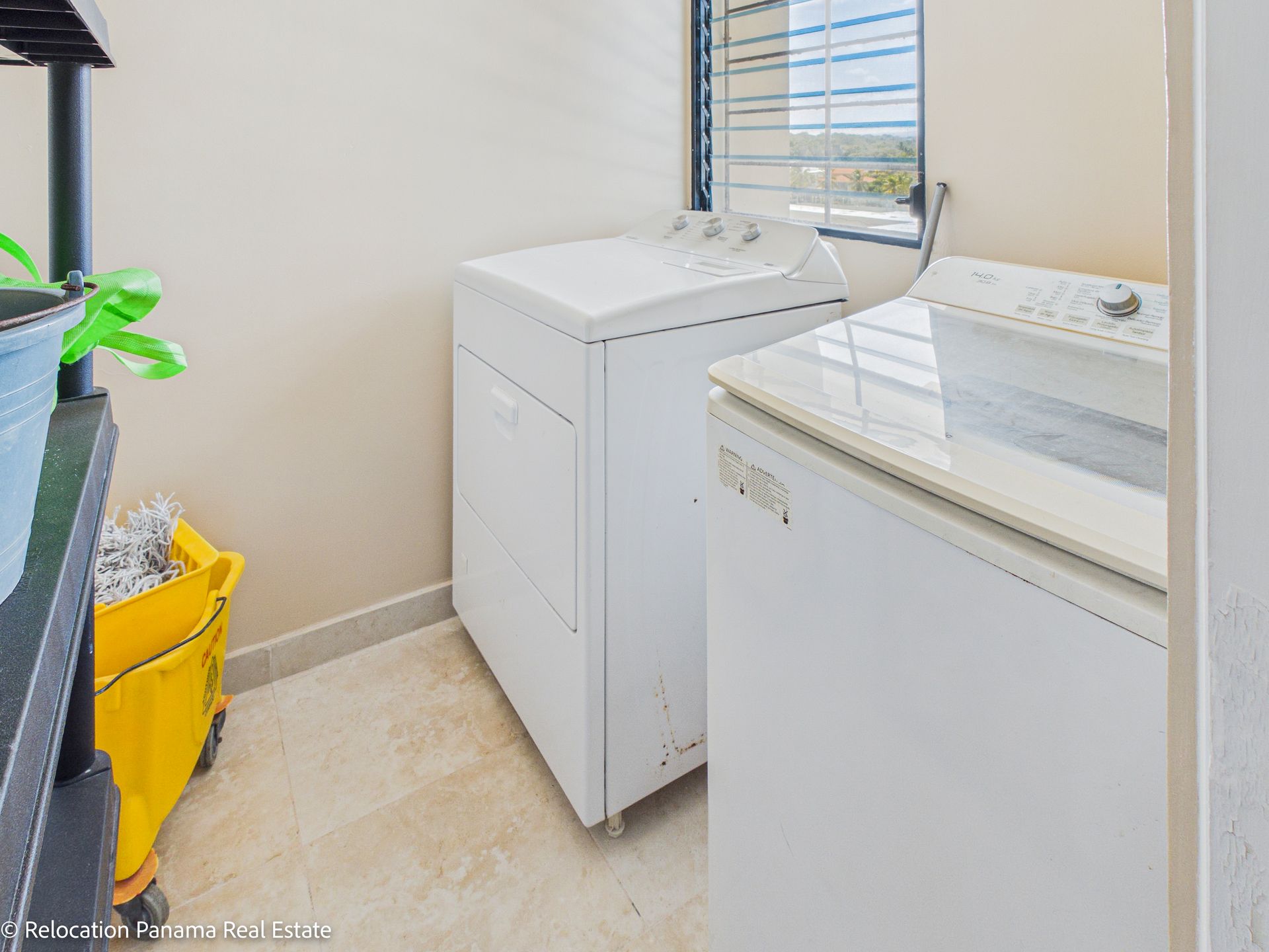 Cuarto de lavandería con dos lavadoras blancas, un carrito de servicio amarillo y una ventana con listones sobre un piso de baldosas.