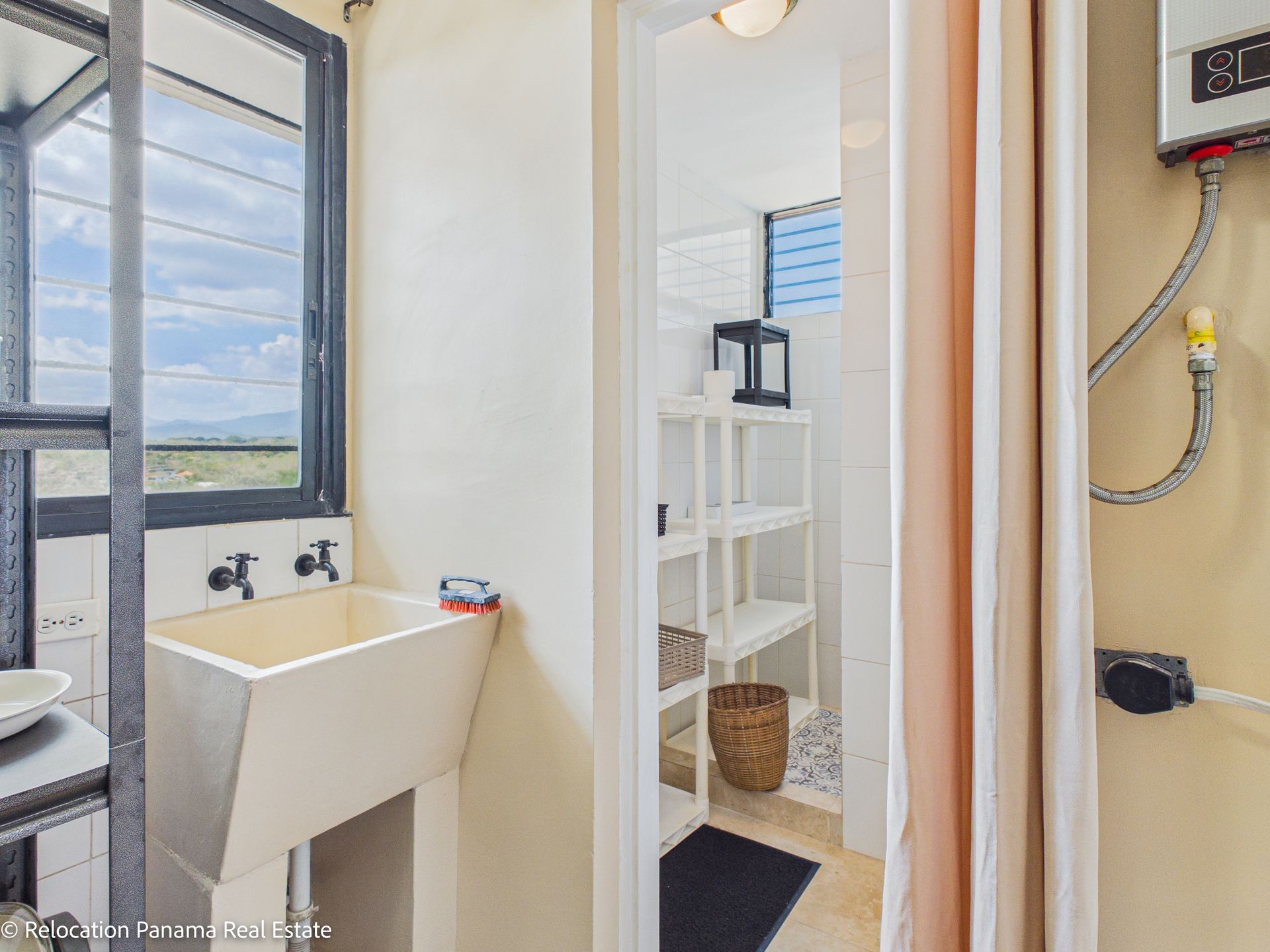 Un lavadero se encuentra junto a una puerta que da a una pequeña habitación alicatada con estanterías blancas y un suelo estampado.