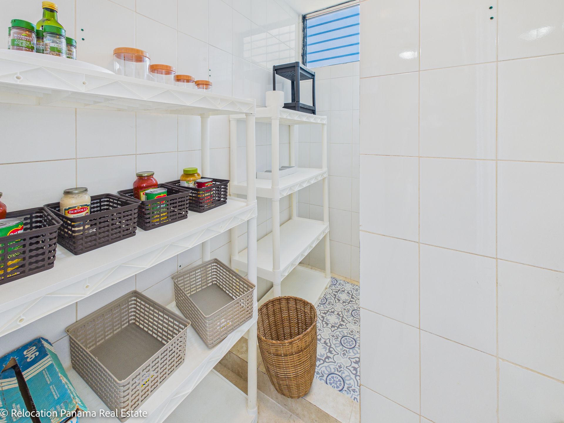 Una despensa con estantes blancos, cestas de plástico para guardar alimentos, varios tarros de cristal y una cesta de mimbre sobre un suelo de baldosas.