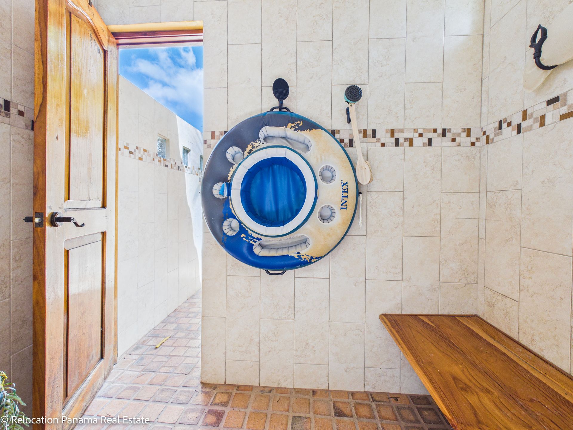 Una zona de ducha al aire libre con paredes alicatadas, un banco de madera, un flotador de piscina azul y blanco colgando y una puerta de madera abierta.