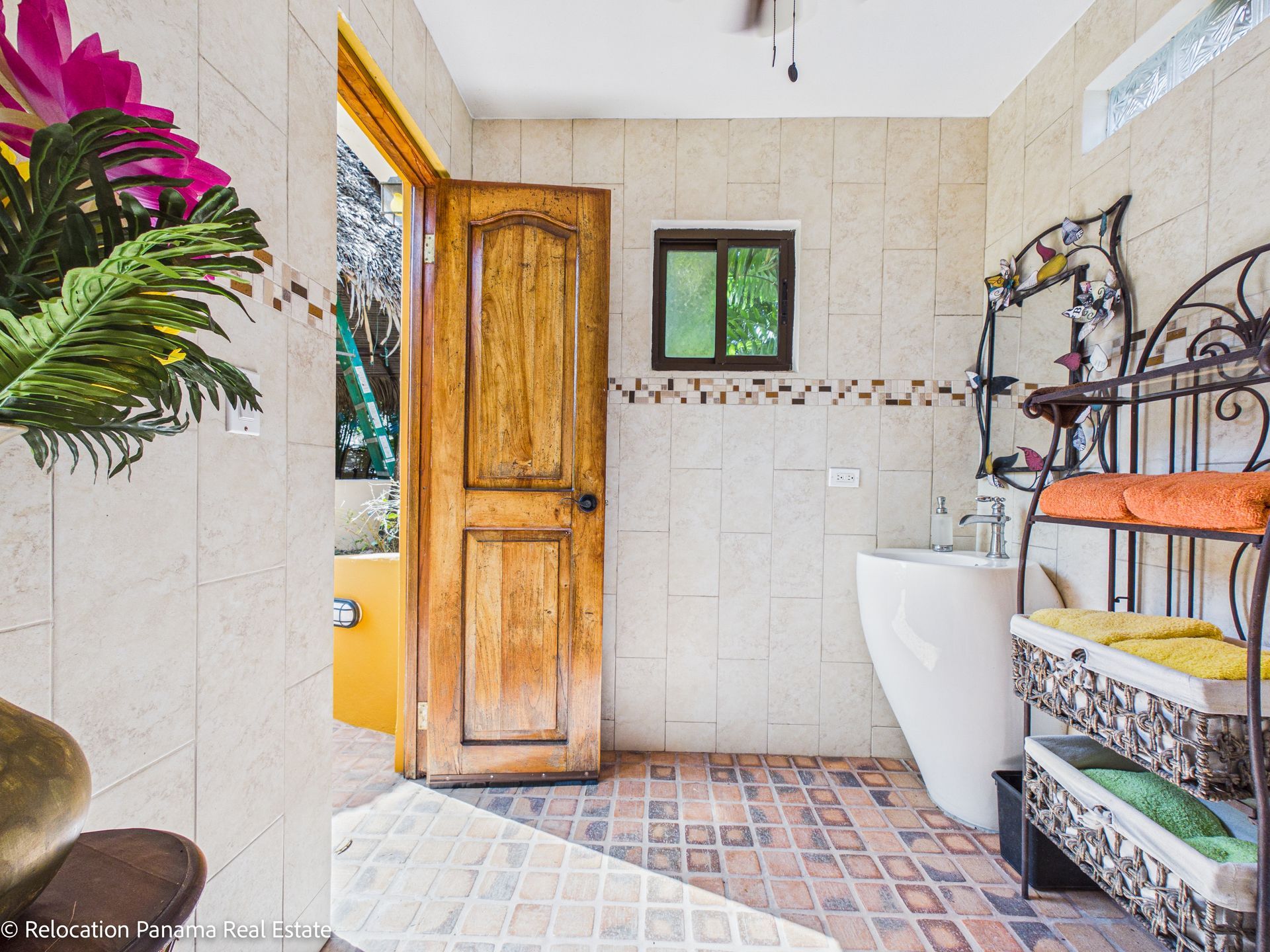 Un baño interior-exterior alicatado con puerta de madera, lavabo blanco con pedestal y estantería metálica con toallas dobladas.