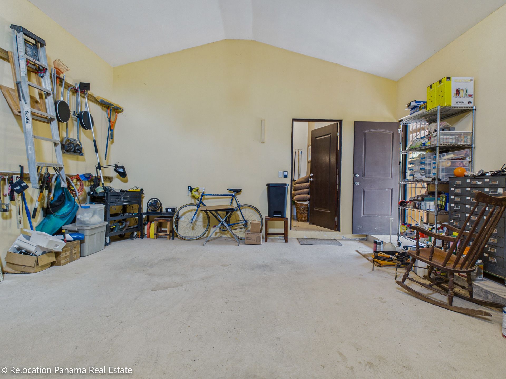 Un garaje austero con paredes amarillas, una bicicleta, una mecedora, herramientas colgadas en la pared y dos puertas abiertas.