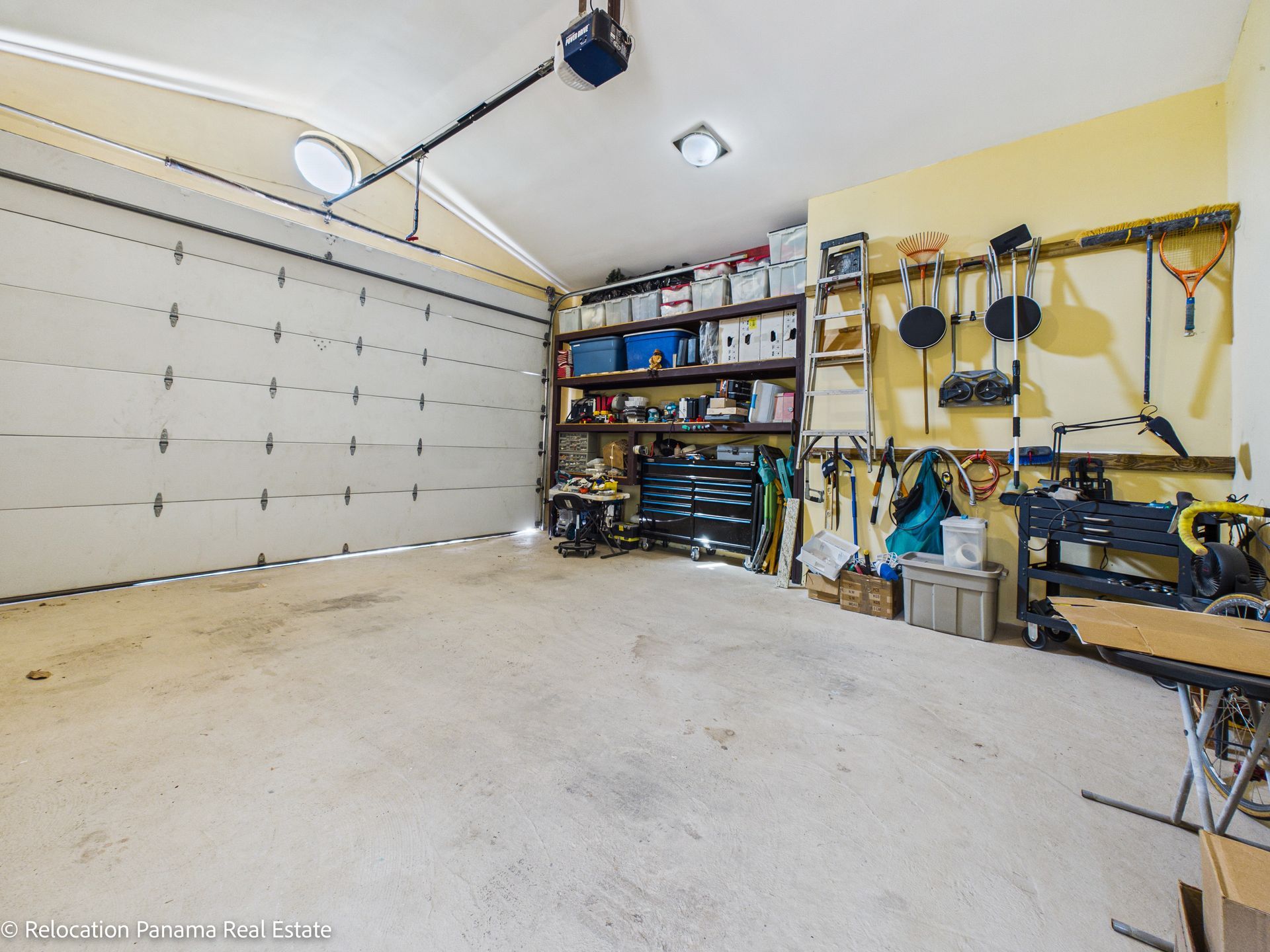 Un garaje espacioso con puerta seccional blanca, estanterías metálicas, espacio para guardar herramientas y equipos colgados en paredes amarillas.