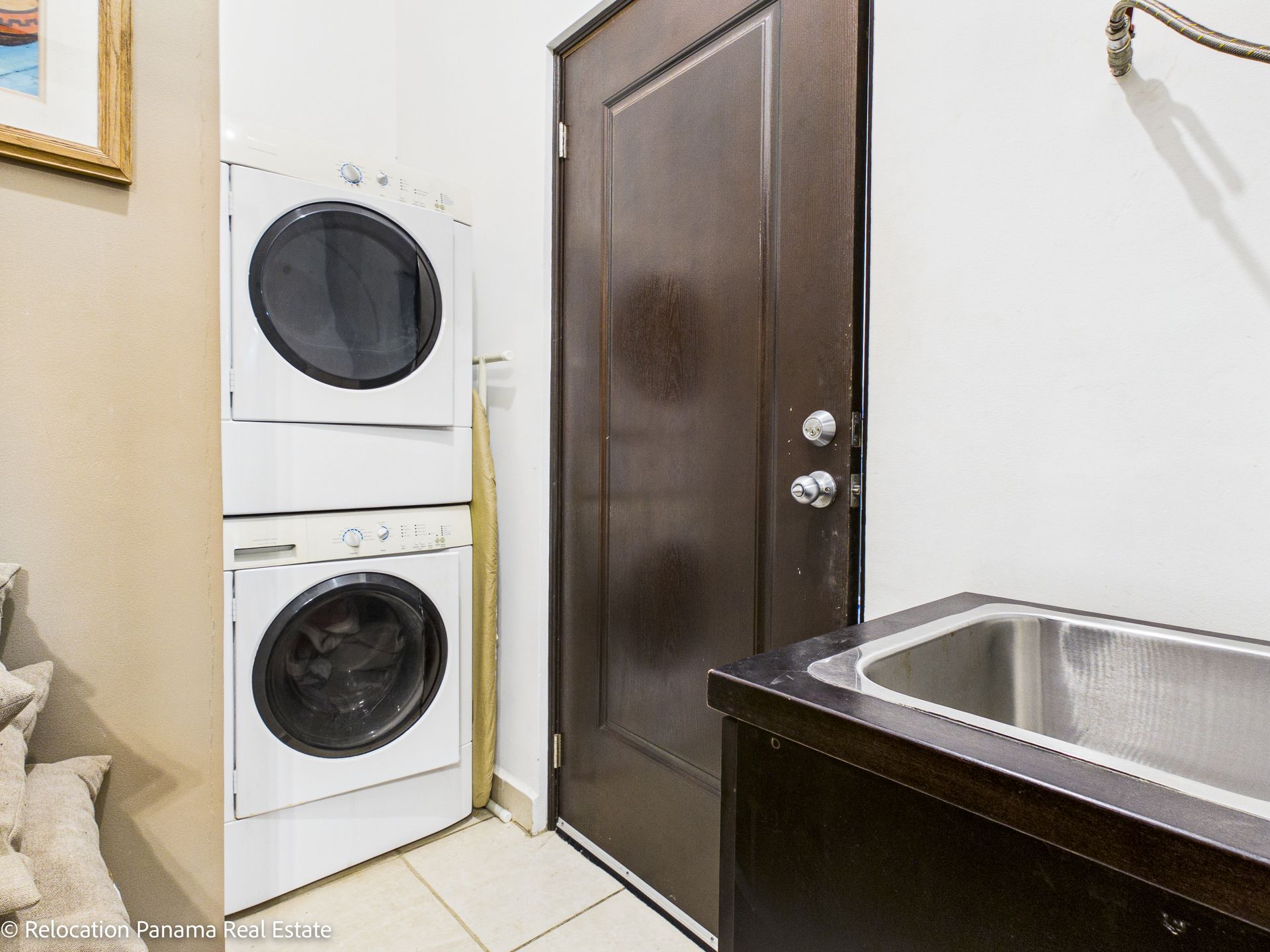 Un área de lavandería con lavadora y secadora apiladas, una puerta de madera oscura y un fregadero de servicio de acero inoxidable.