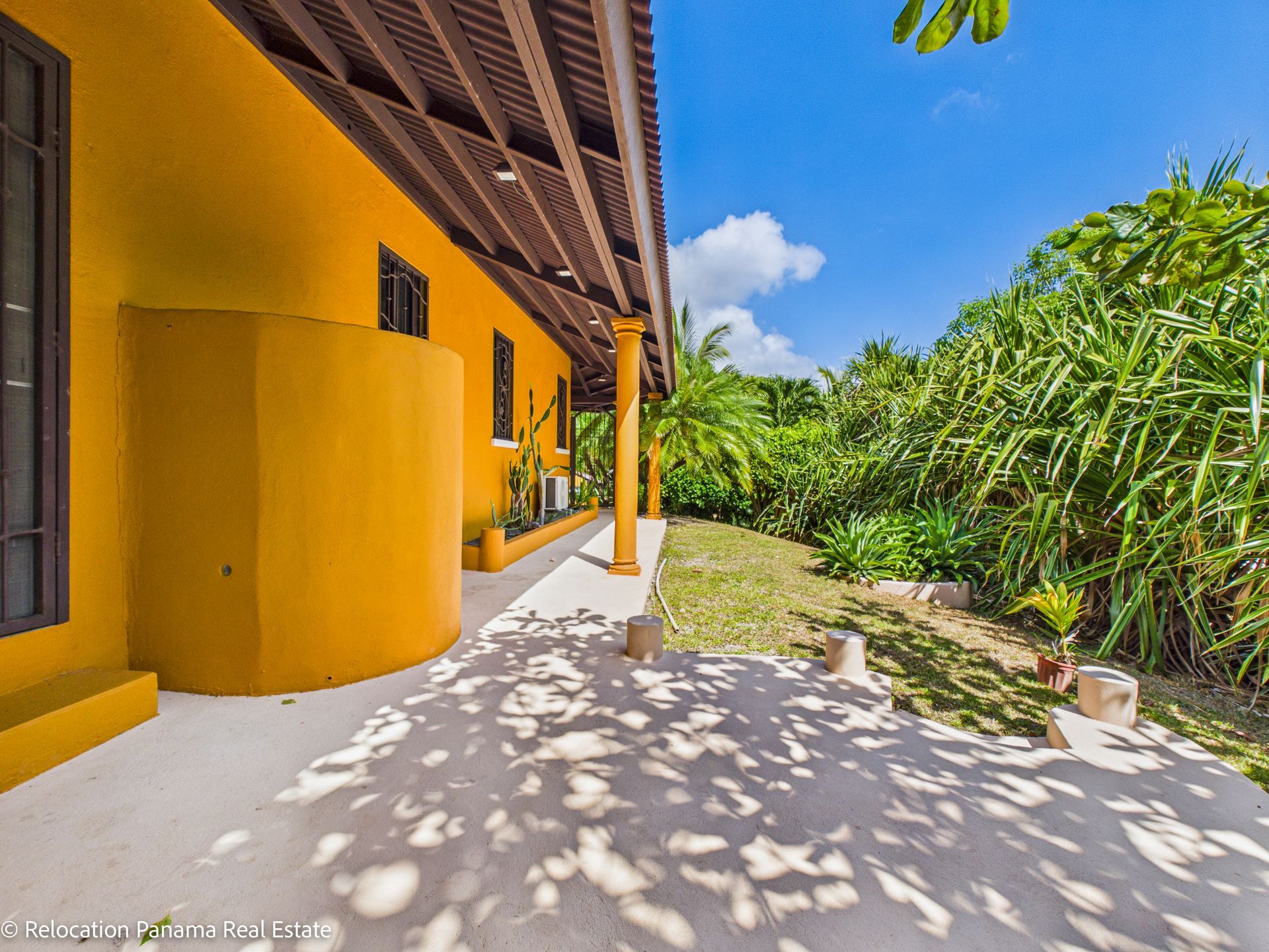 Un patio junto a una casa de color amarillo brillante con techo cubierto, rodeado de exuberante vegetación verde bajo un cielo azul soleado.