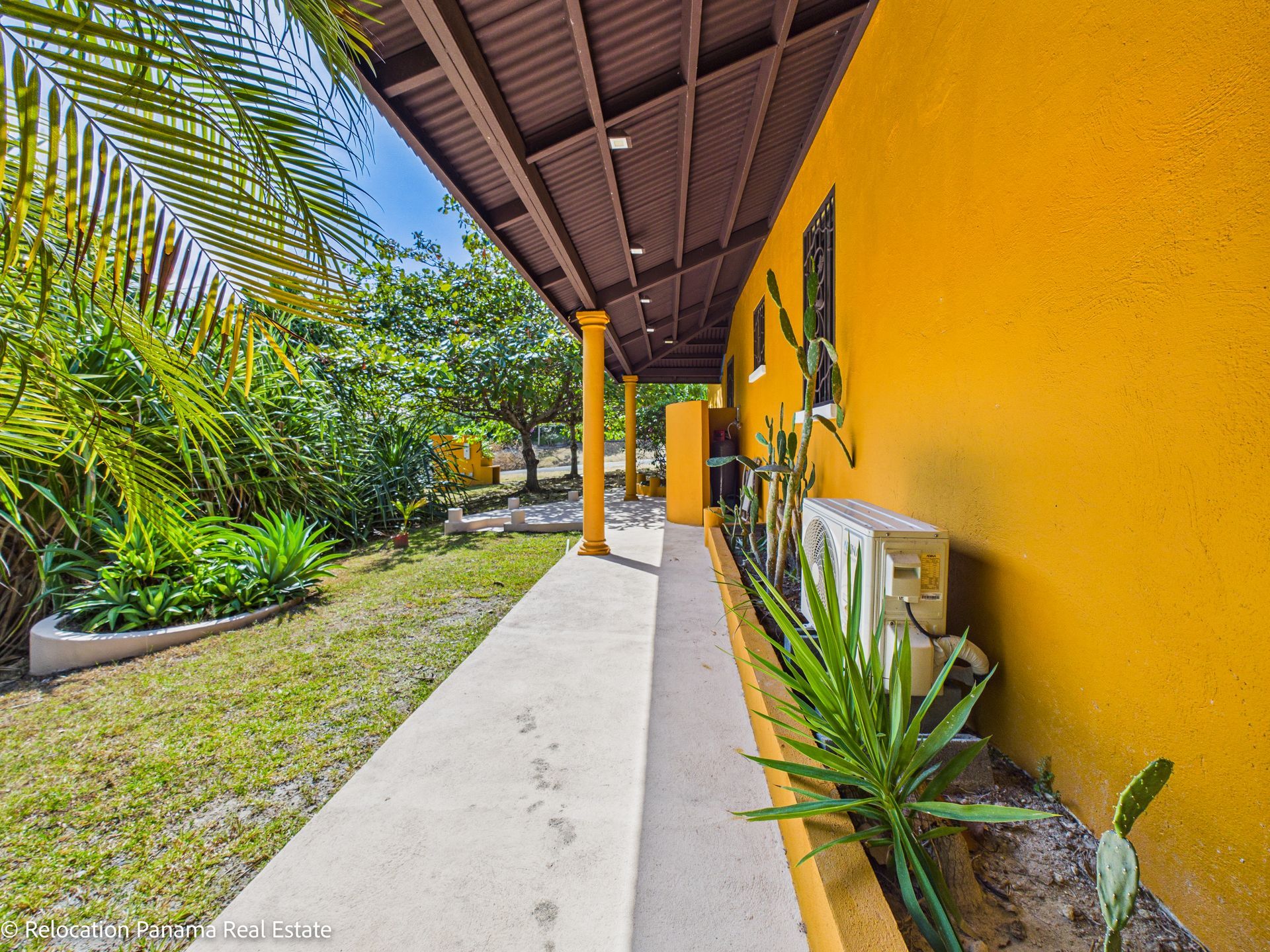 El exterior de una casa amarilla con una pasarela cubierta y un patio junto a un jardín con césped, plantas y árboles.