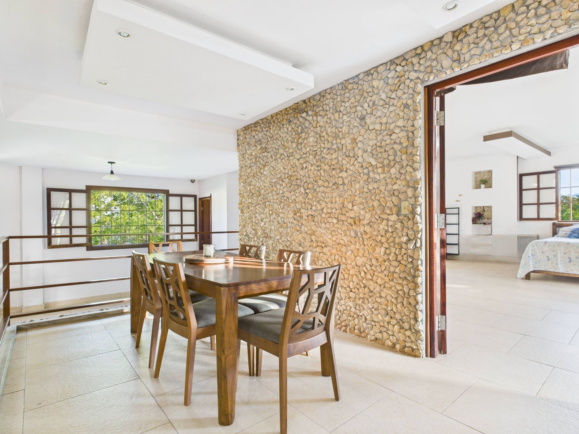 Comedor con mesa y sillas de madera, pared de piedra y puerta de acceso a un dormitorio con cama.