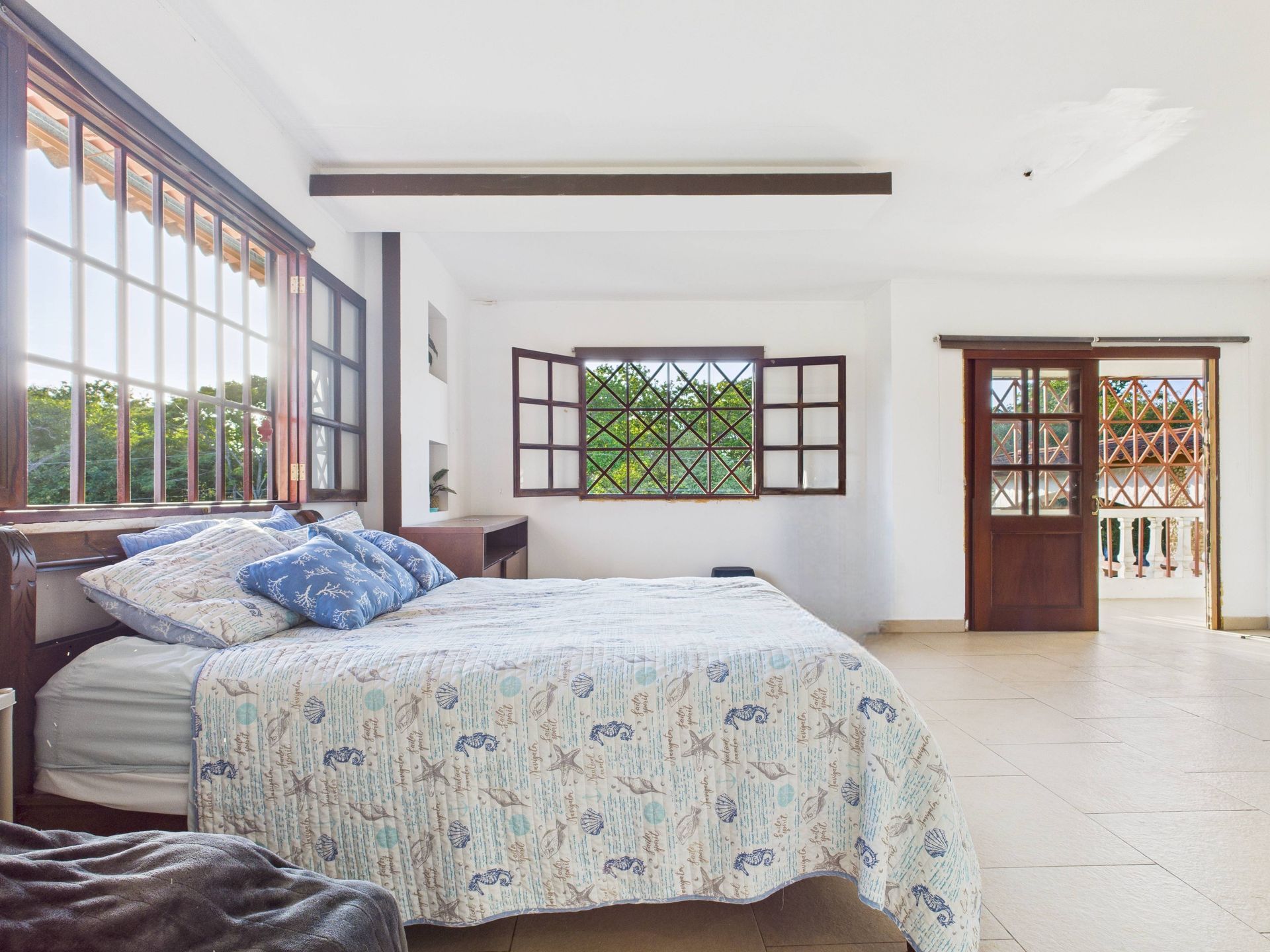 Dormitorio con grandes ventanales, cama de matrimonio, contraventanas de madera y puerta que da a balcón.