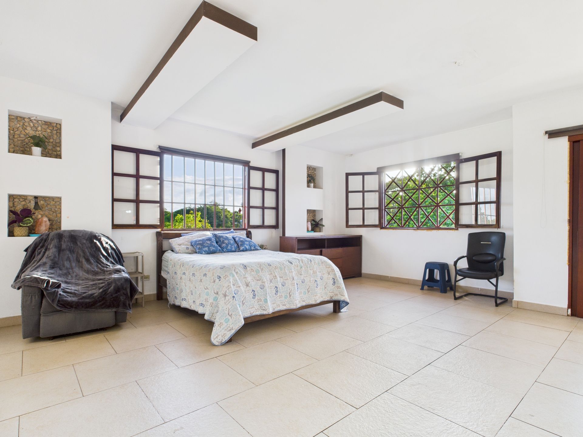 Amplio dormitorio con cama, ventanas y asientos. Tonos neutros y suelo de baldosas claras.