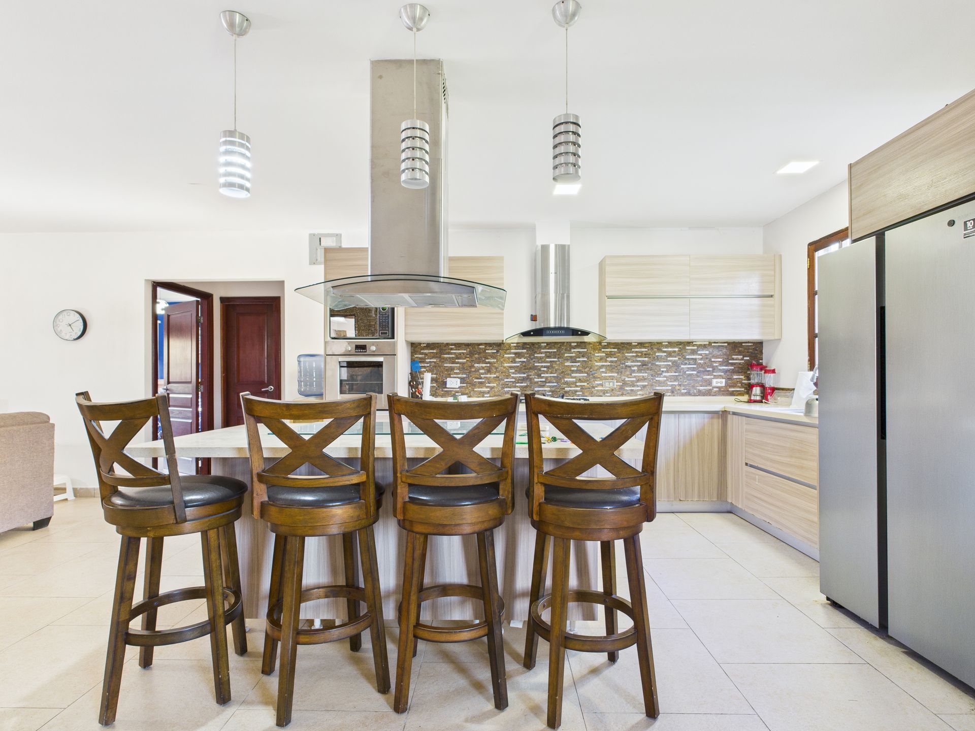 Cocina con isla de asientos, gabinetes de madera clara, electrodomésticos de acero inoxidable e iluminación colgante.