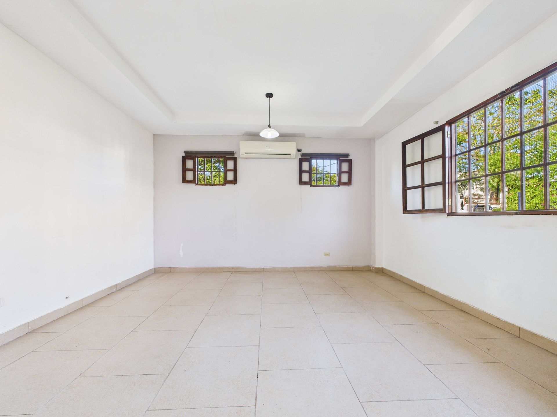 Habitación vacía con paredes blancas, suelo de baldosas, ventanas y unidad de aire acondicionado.