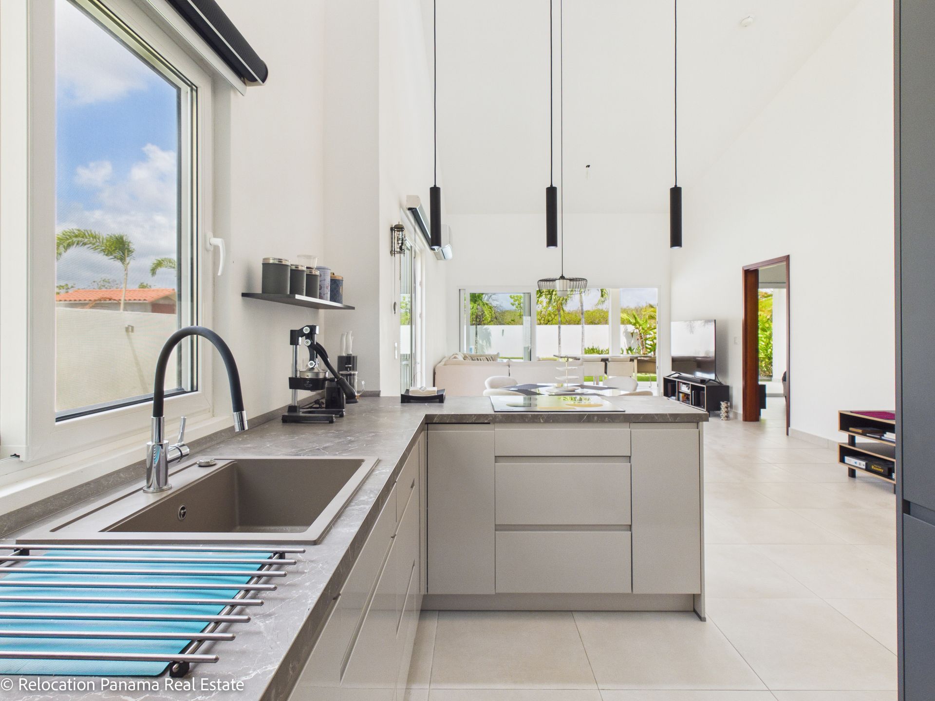 Cocina moderna con armarios beige, encimera oscura, fregadero junto a la ventana y tres lámparas colgantes en un espacio diáfano.