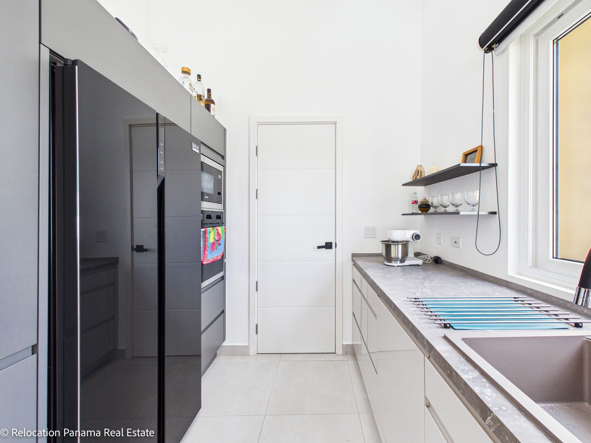 Una cocina moderna y minimalista cuenta con armarios grises, un frigorífico oscuro, paredes blancas y una larga encimera con fregadero.