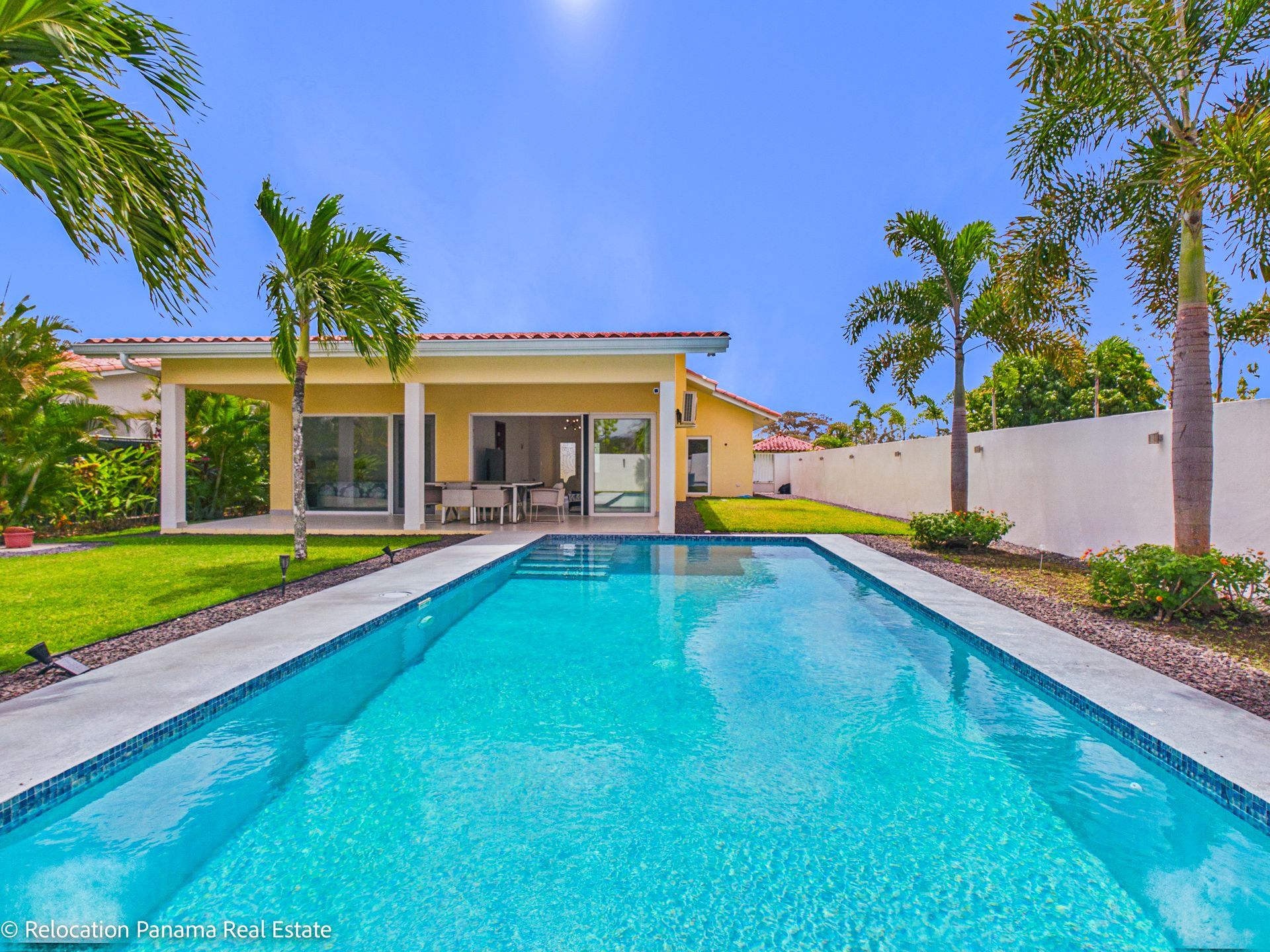Un patio trasero soleado con una piscina rectangular, palmeras, césped verde y una casa amarilla con un patio cubierto.