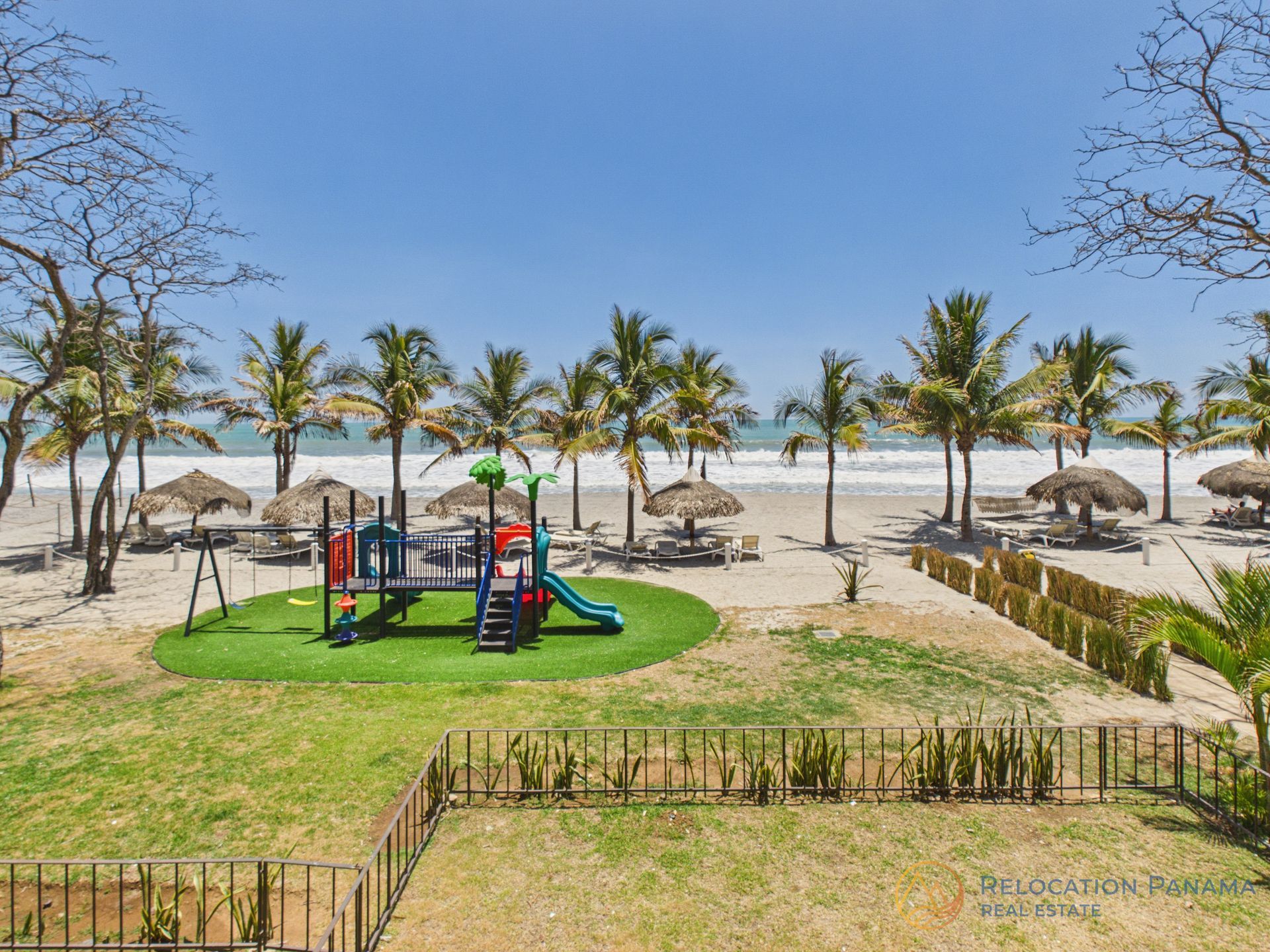 Un parque infantil con tobogán en una zona de césped junto a una playa de arena, bordeada de palmeras y sombrillas con vistas al mar.