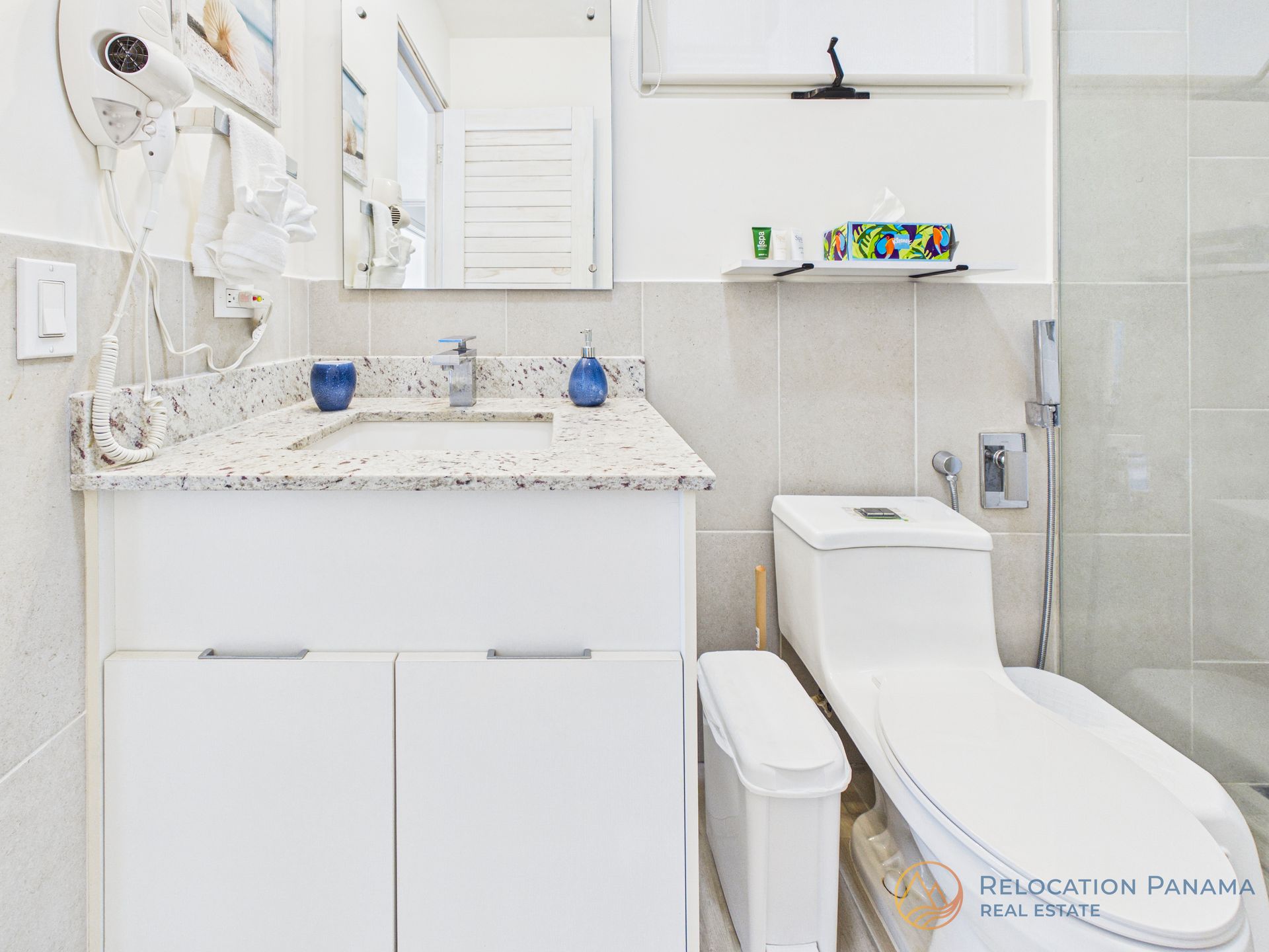 Un baño con un tocador blanco con encimera moteada, un inodoro, una papelera estrecha y una ducha con mampara de cristal.