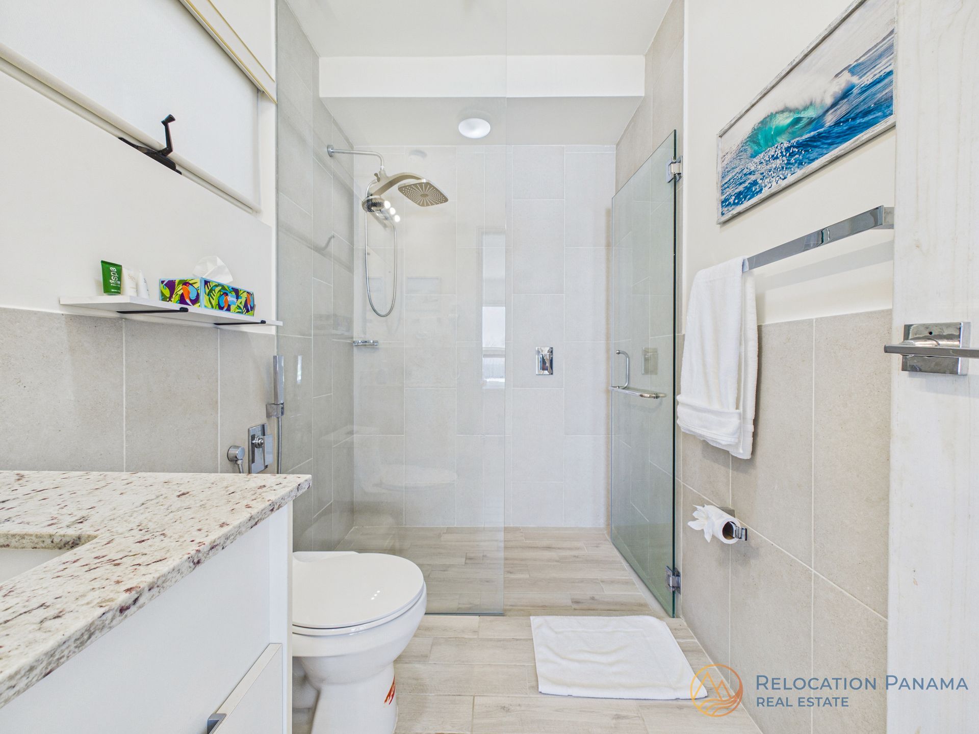 Un baño moderno con tocador con encimera de granito, inodoro blanco y ducha a ras de suelo con mampara de cristal y paredes alicatadas.