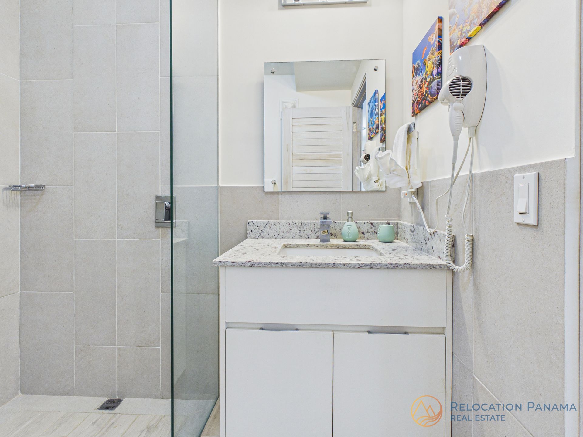 Un moderno baño de hotel con ducha acristalada, tocador blanco con encimera de granito y secador de pelo.