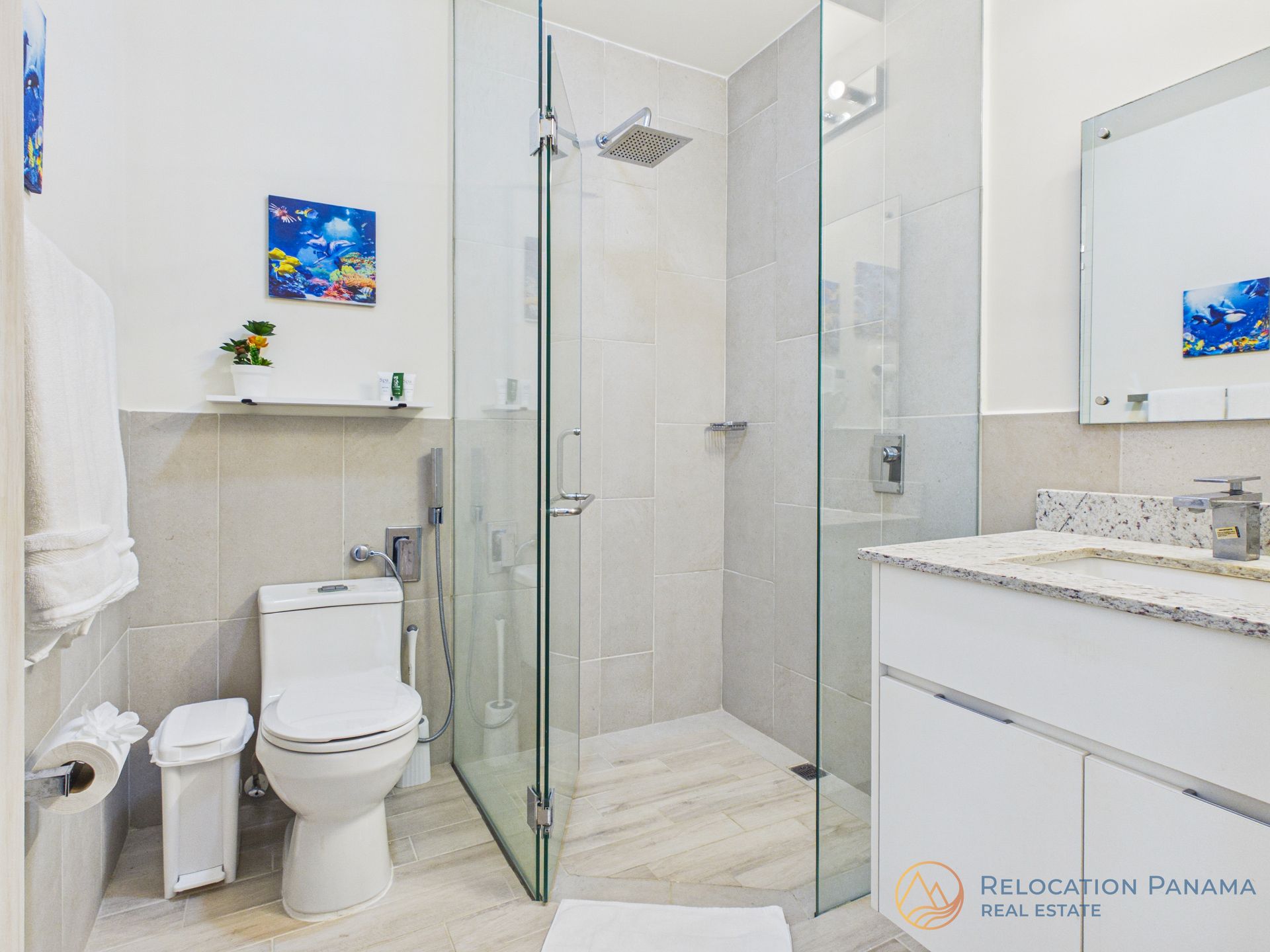 Un baño moderno con inodoro blanco, ducha con mampara de cristal, tocador con encimera de granito y obras de arte decorativas en la pared.