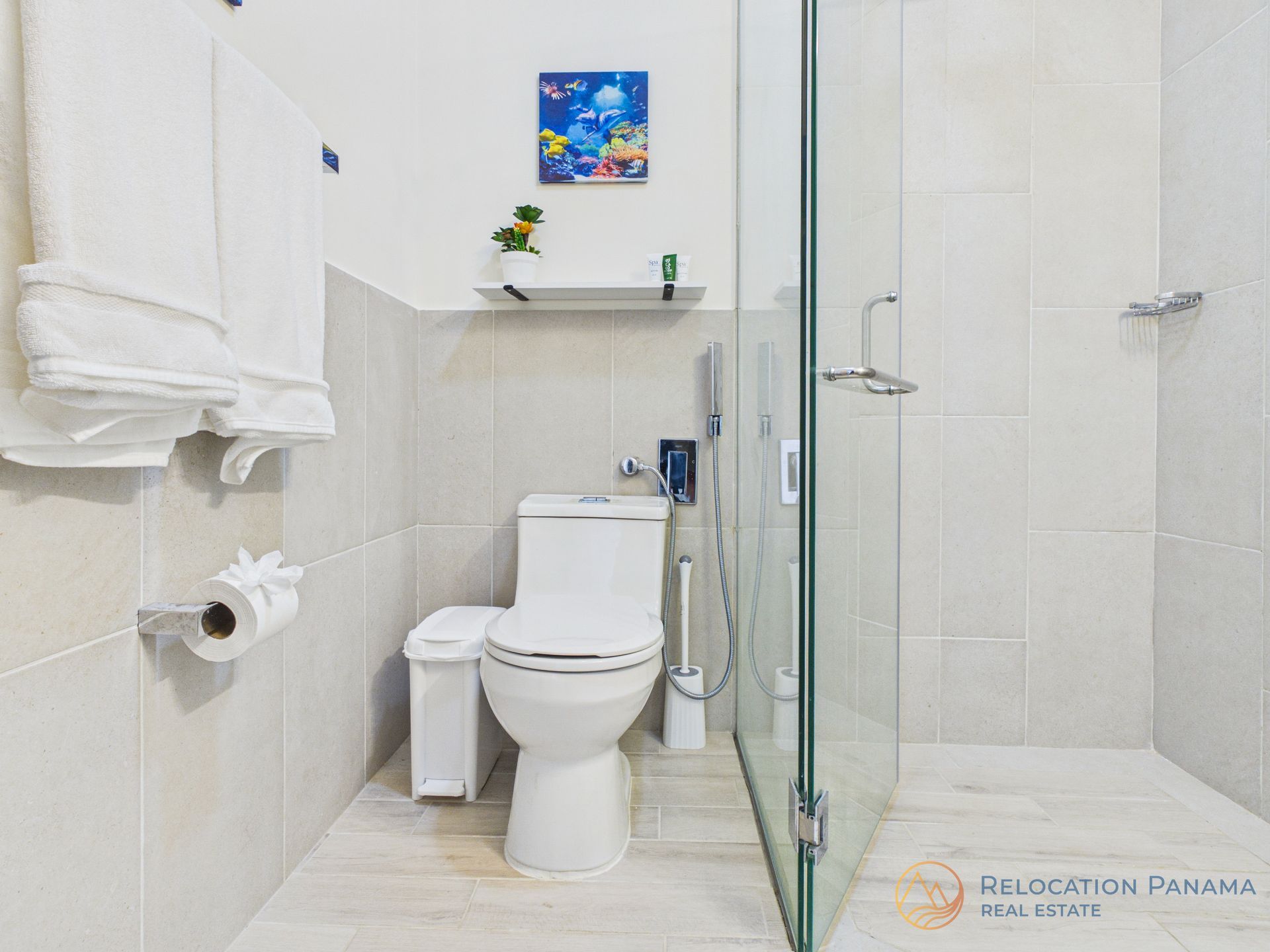 Un baño moderno con inodoro blanco, ducha con mampara de cristal, estante de pared y toallas colgadas en la pared.