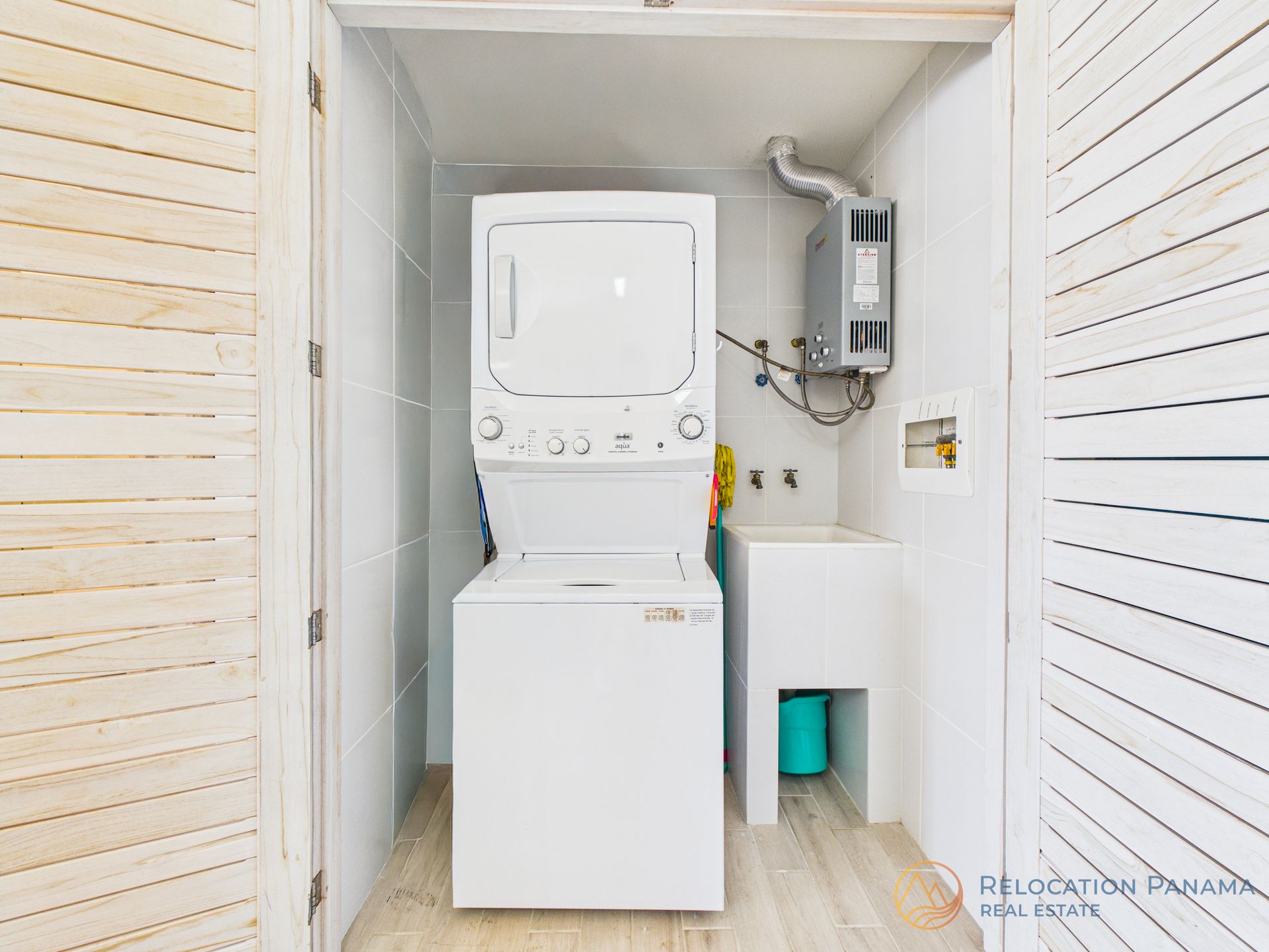 Un cuarto de lavandería con una lavadora y secadora apiladas de color blanco, un fregadero blanco y un calentador de agua mural.