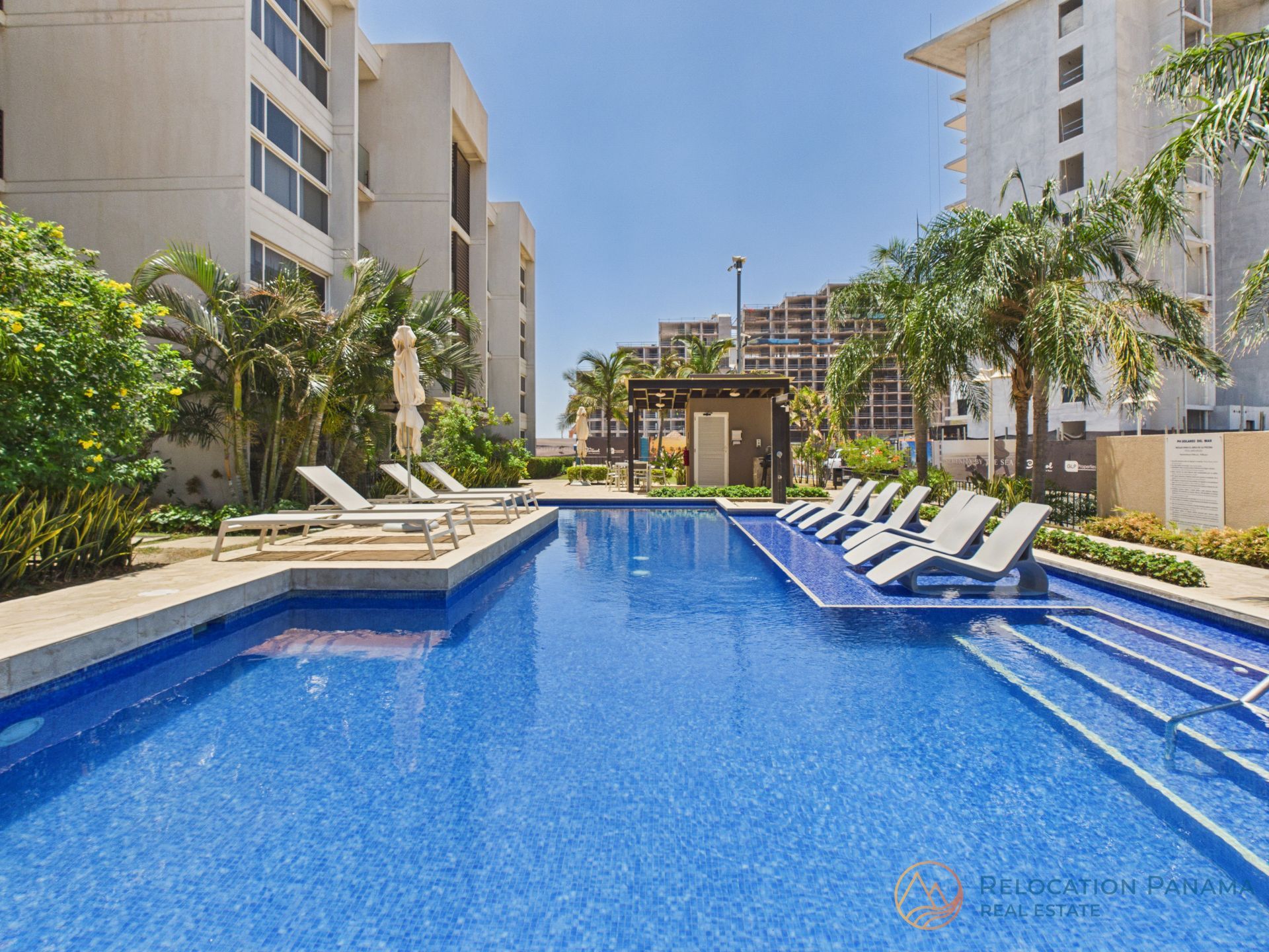 Una piscina exterior de color azul brillante rodeada de tumbonas y modernos edificios de apartamentos de varias plantas. playa caracol chame