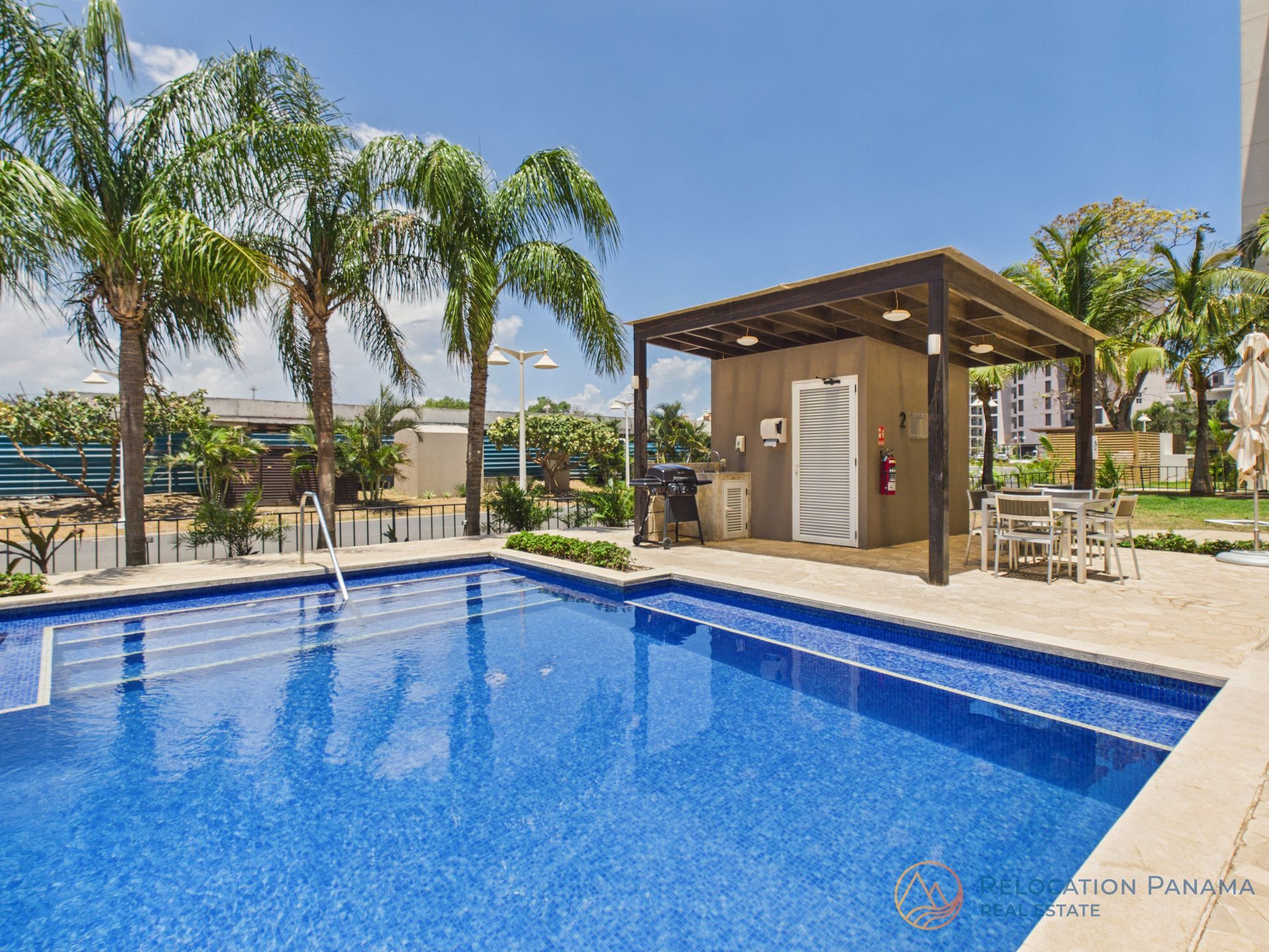 Una vibrante piscina azul se ubica junto a una pérgola de madera con muebles de exterior para comer, en un día soleado rodeado de palmeras. playa caracol chame