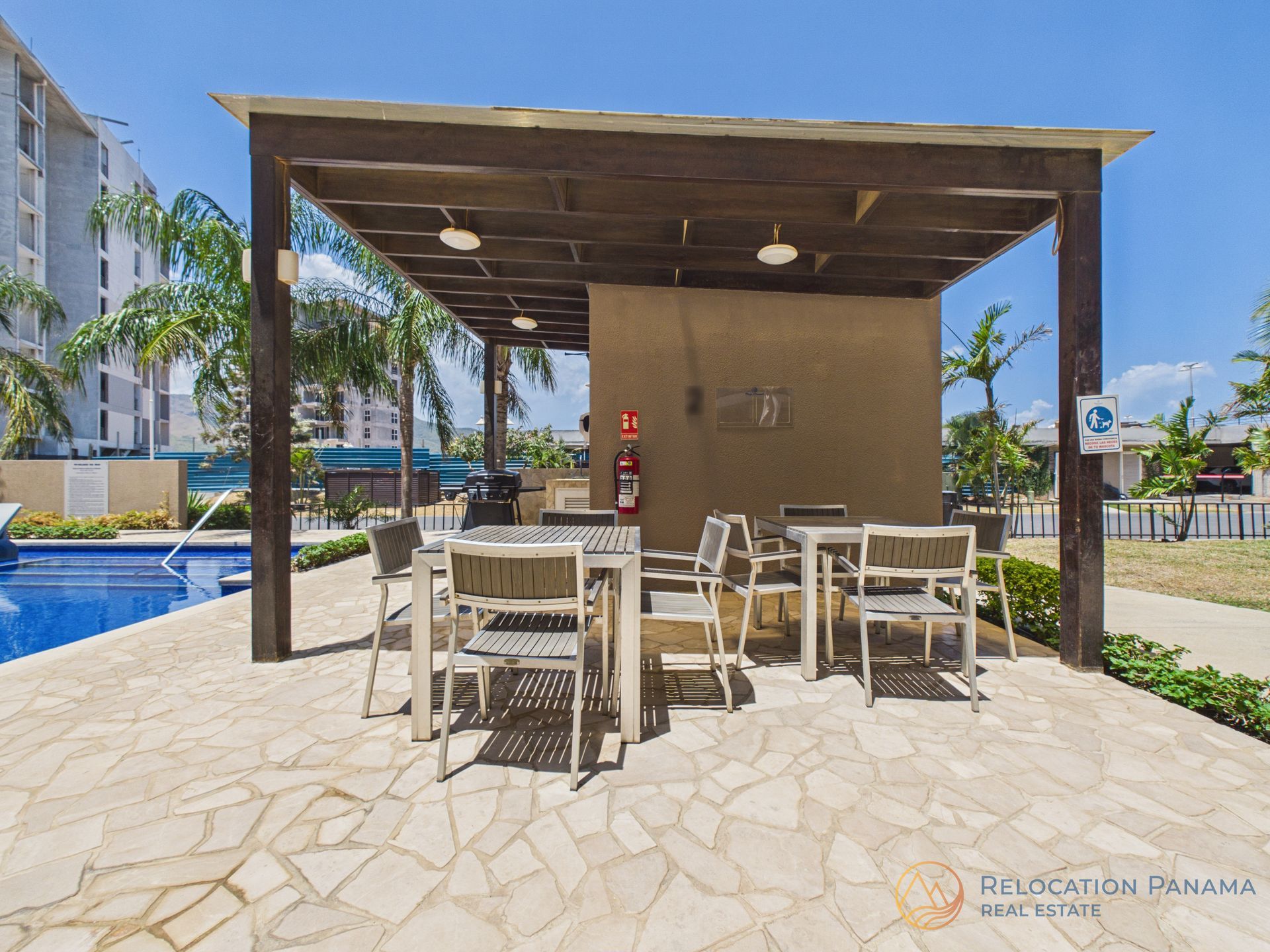 Patio exterior con mesas y sillas de comedor bajo una pérgola de madera junto a una piscina en un día soleado. playa caracol chame