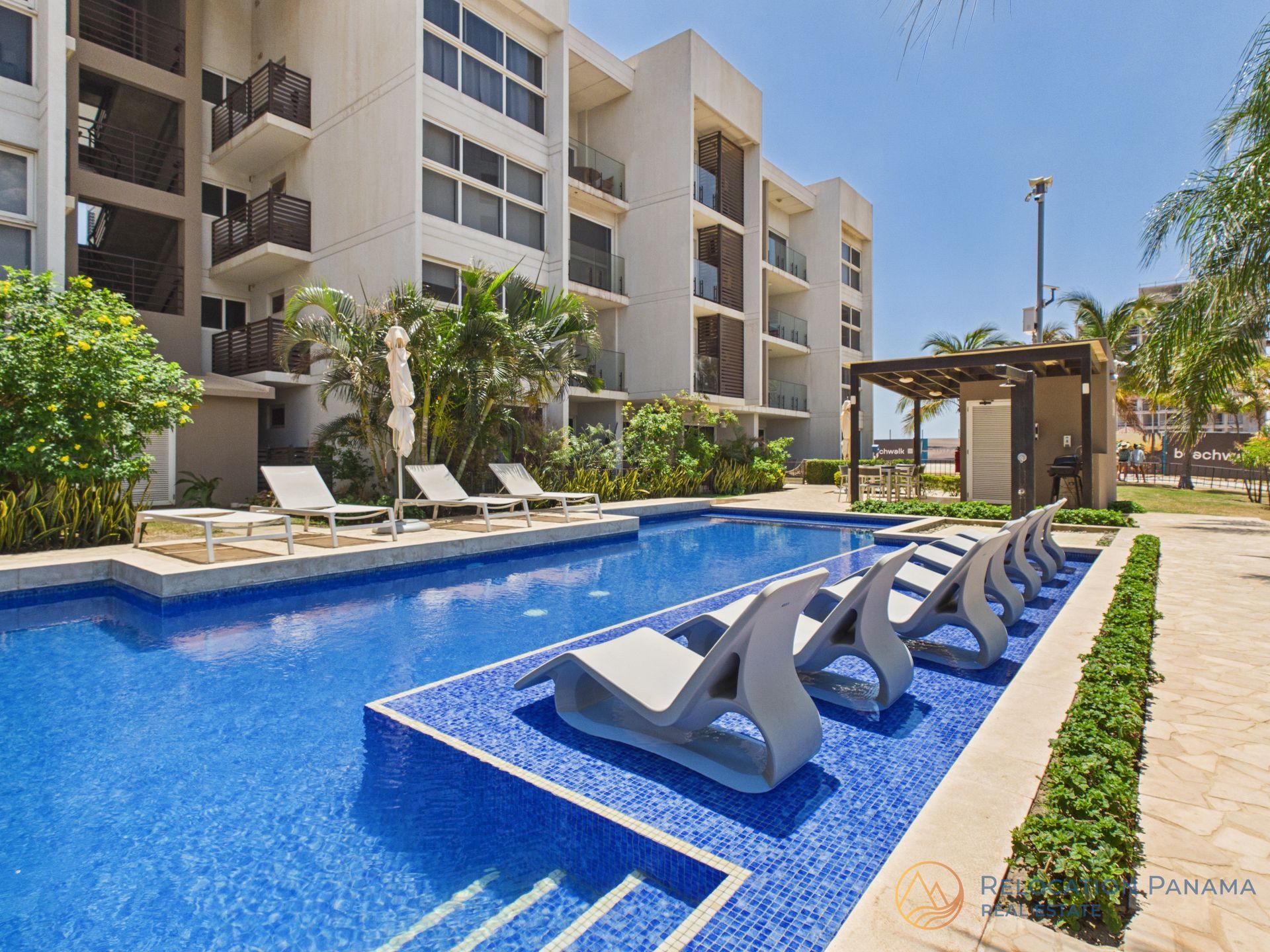 Una piscina con tumbonas parcialmente sumergidas en aguas poco profundas, situada junto a un moderno edificio de apartamentos de varias plantas. playa caracol chame