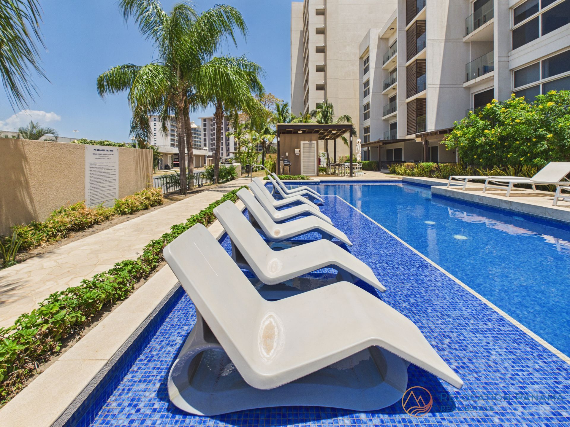 Tumbonas de color beige sumergidas en una piscina con azulejos azules, junto a un edificio moderno y palmeras. playa caracol chame