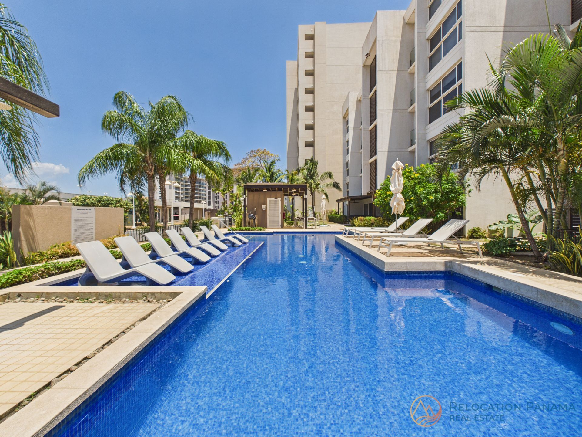 Una larga piscina con azulejos azules, rodeada de tumbonas, palmeras y un alto y moderno edificio de apartamentos. playa caracol chame