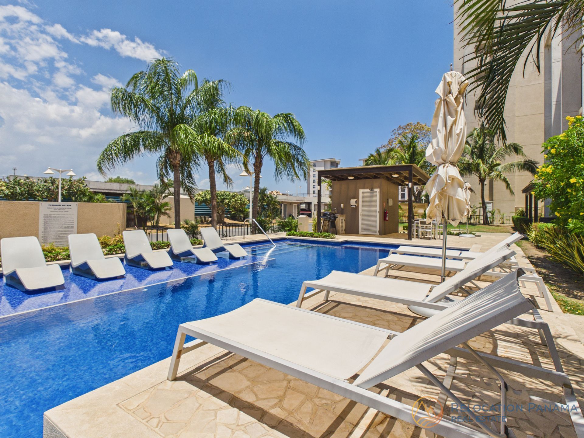 Una piscina rectangular azul rodeada de tumbonas blancas, palmeras y un edificio beige bajo un cielo soleado. playa caracol chame