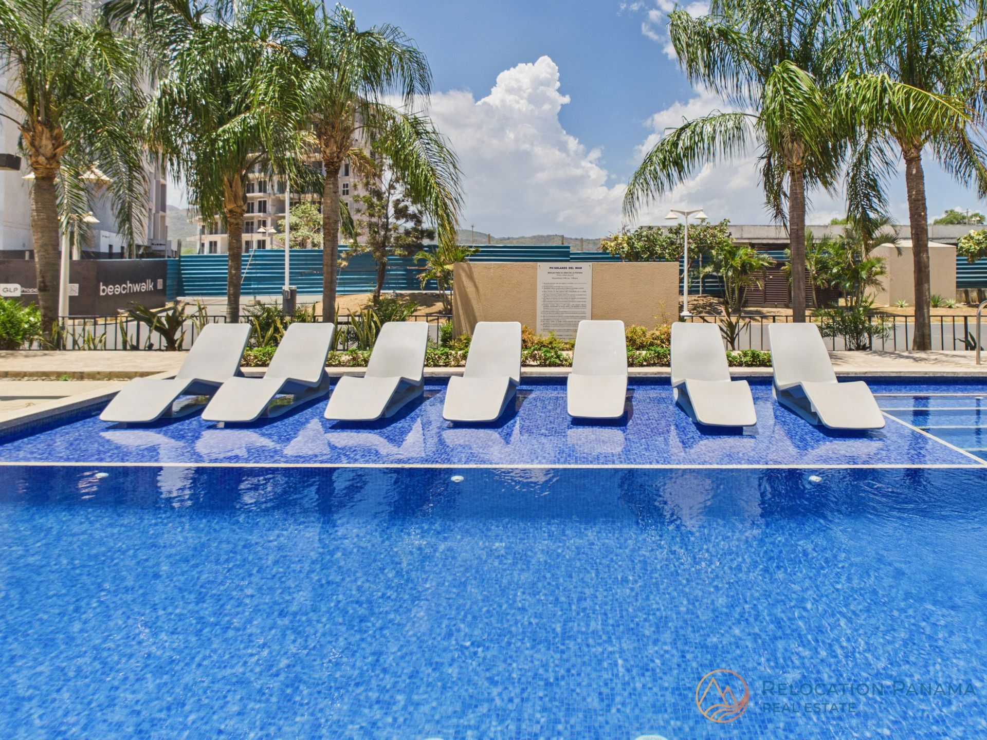 Siete tumbonas blancas colocadas sobre una plataforma sumergida para tomar el sol en una piscina de color azul brillante bajo un cielo soleado. playa caracol chame
