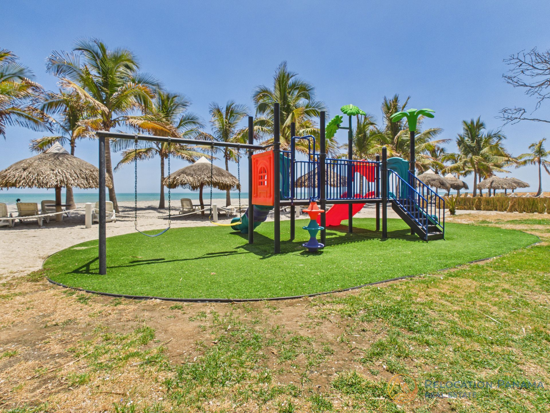 Un colorido parque infantil situado sobre un trozo de césped artificial cerca de una playa soleada con palmeras y sombrillas de paja.playa caracol chame