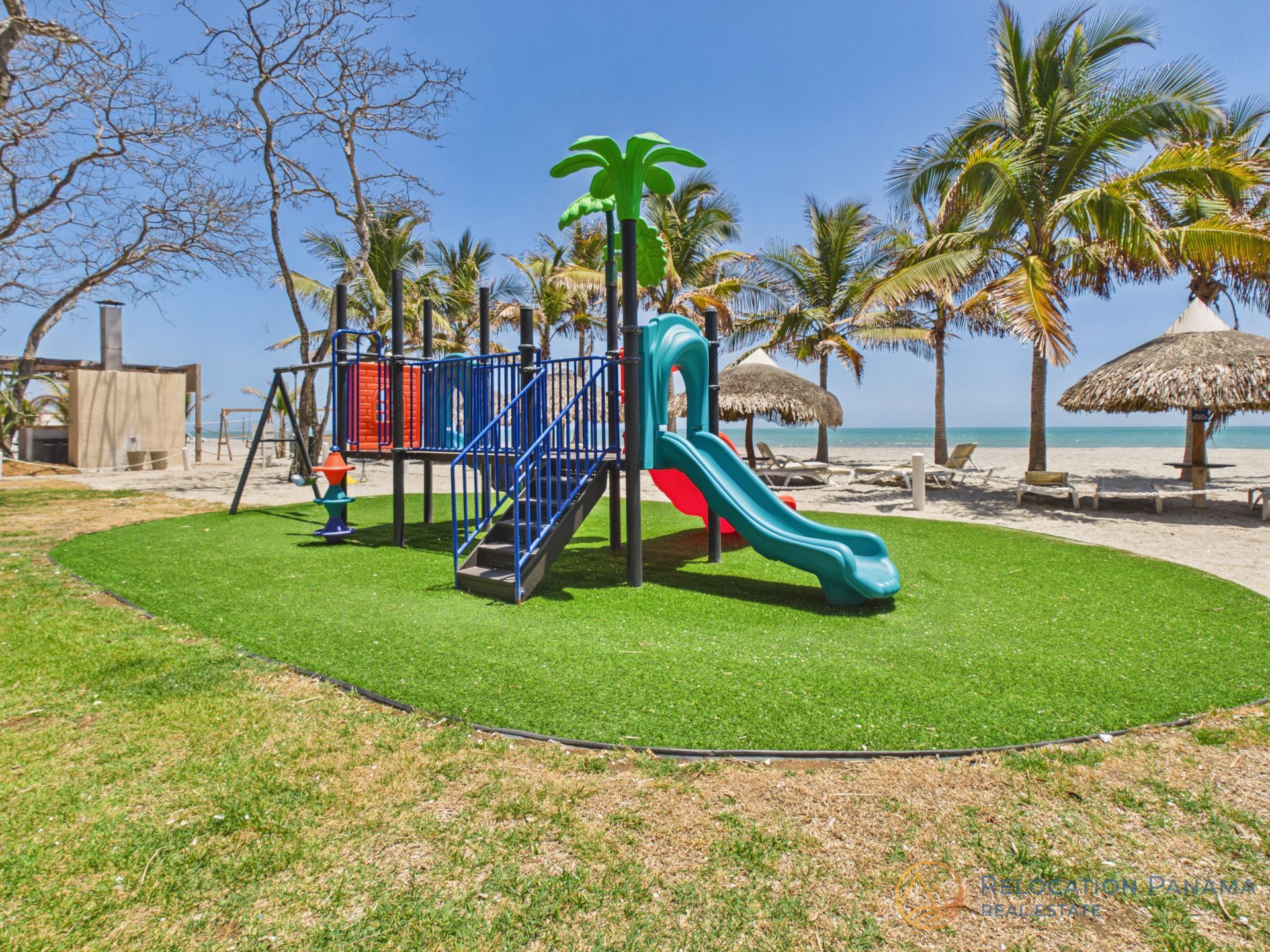 Un colorido parque infantil situado en una zona de césped junto a una playa de arena con palmeras y sombrillas. playa caracol chame