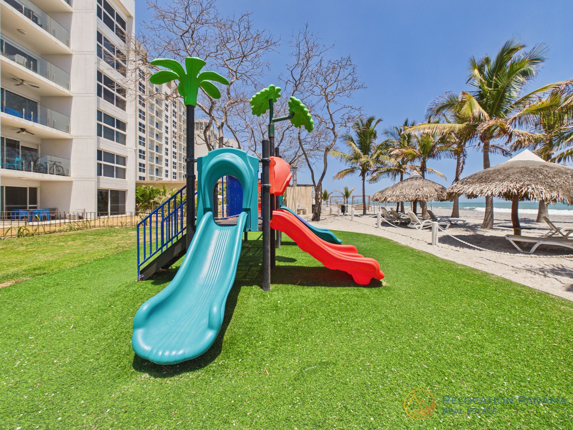 Un colorido tobogán infantil situado en un césped junto a un alto edificio residencial y una playa soleada con palmeras. playa caracol chame