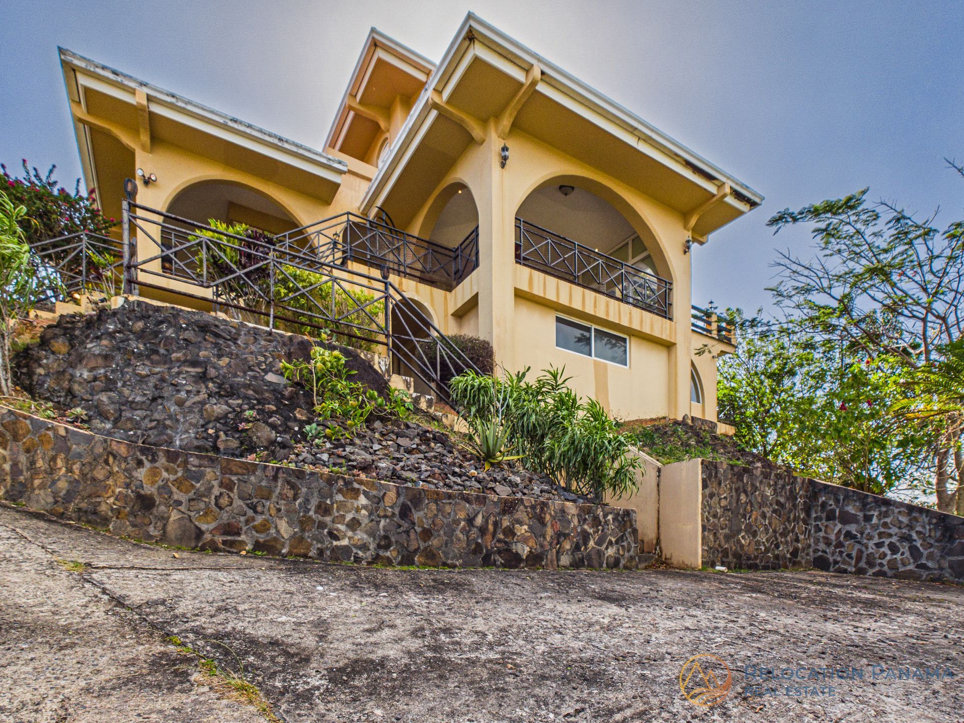Una casa amarilla de dos plantas con porches arqueados, construida sobre una ladera rocosa escalonada, con un cielo azul brillante de fondo.