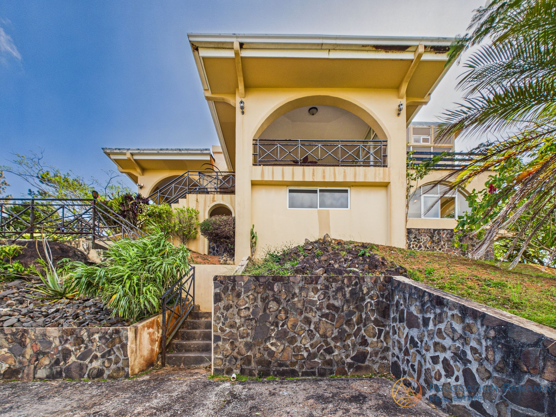 Una casa amarilla de dos plantas con balcón arqueado, elevada sobre una base de piedra, situada entre una exuberante vegetación bajo un cielo azul.