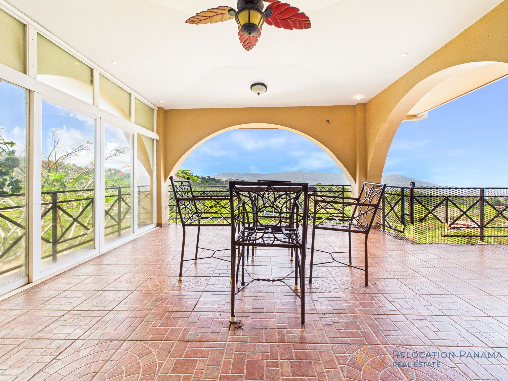 Un patio exterior cubierto con suelo de baldosas, un conjunto de comedor de hierro, ventilador de techo y una abertura arqueada con vistas al paisaje.