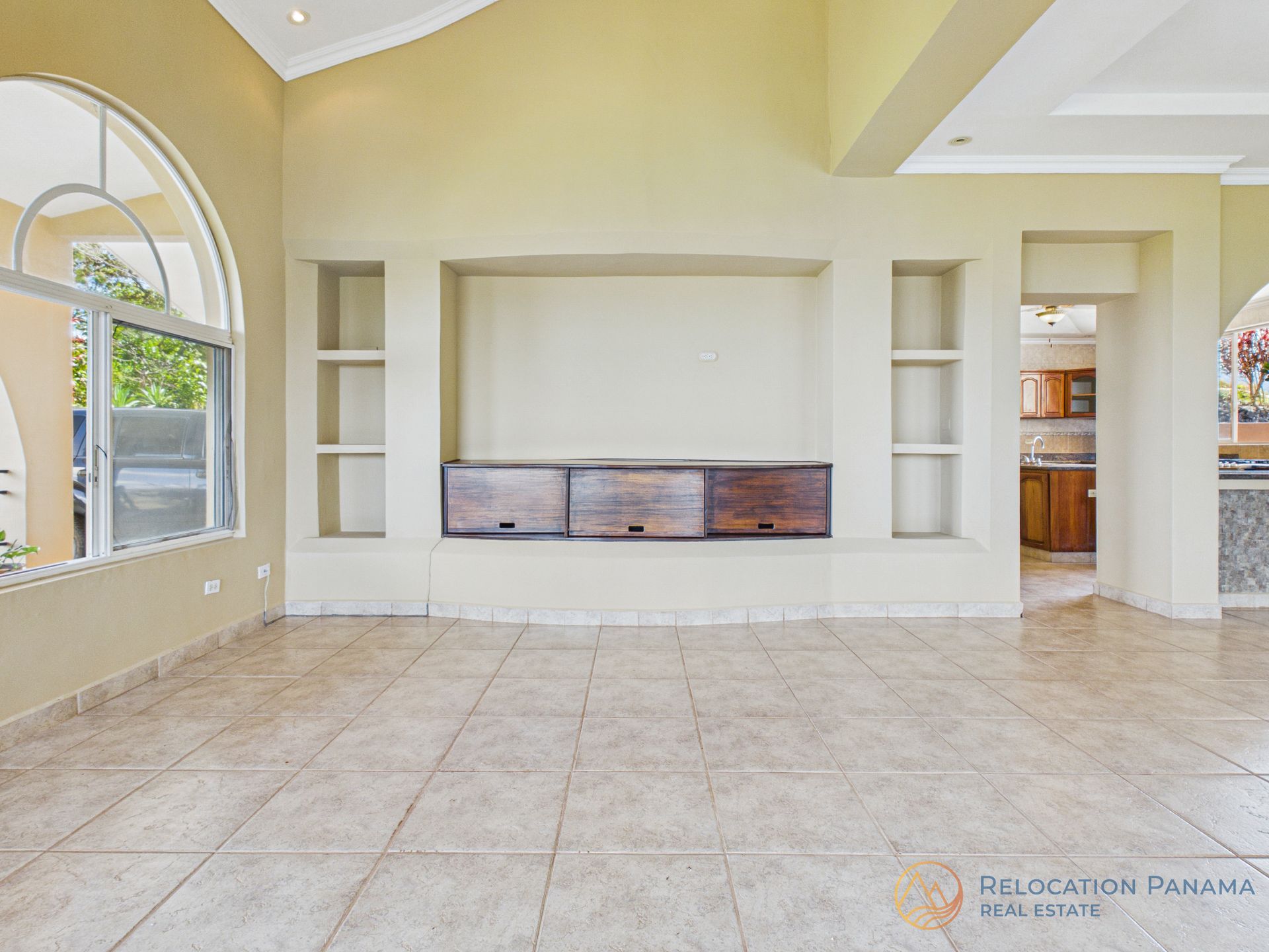 Una habitación espaciosa con paredes de color beige, suelo de baldosas, un gran ventanal arqueado, un rincón multimedia integrado y una puerta que da a la cocina.